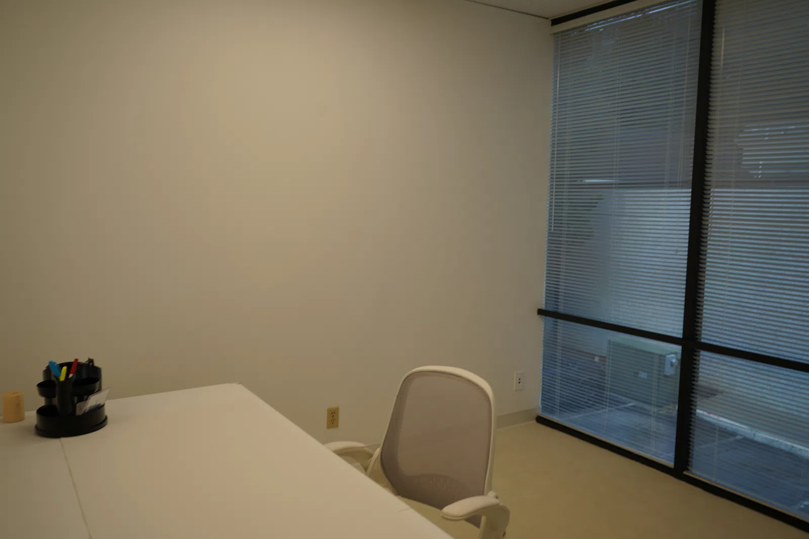 Minimalist office with white desk, ergonomic chair, pen holder, and large windows with blinds.