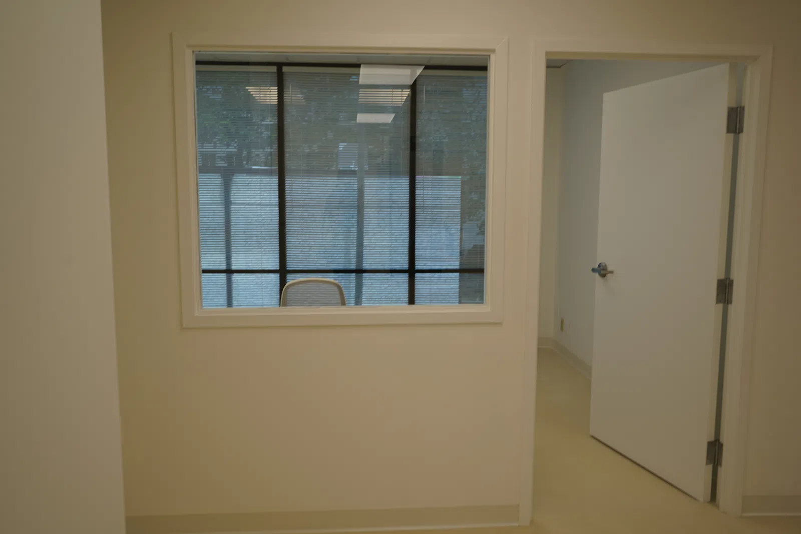 Empty office space with a glass window showing a chair and an open door leading to another room.