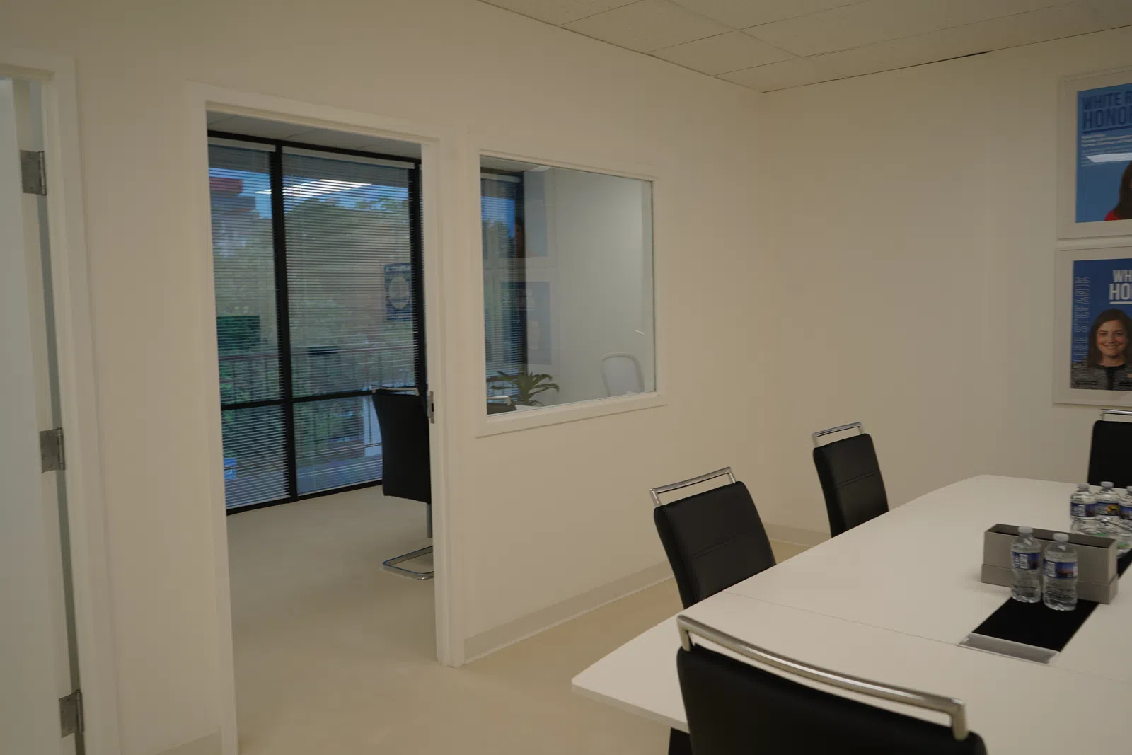 Modern office meeting room with black chairs, white table, water bottles, and glass window to adjacent room.