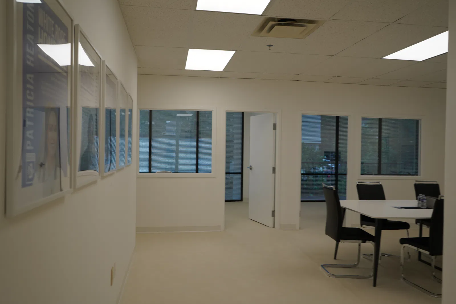 Bright office space with white walls, framed pictures, windows, and a small meeting area with black chairs and table.