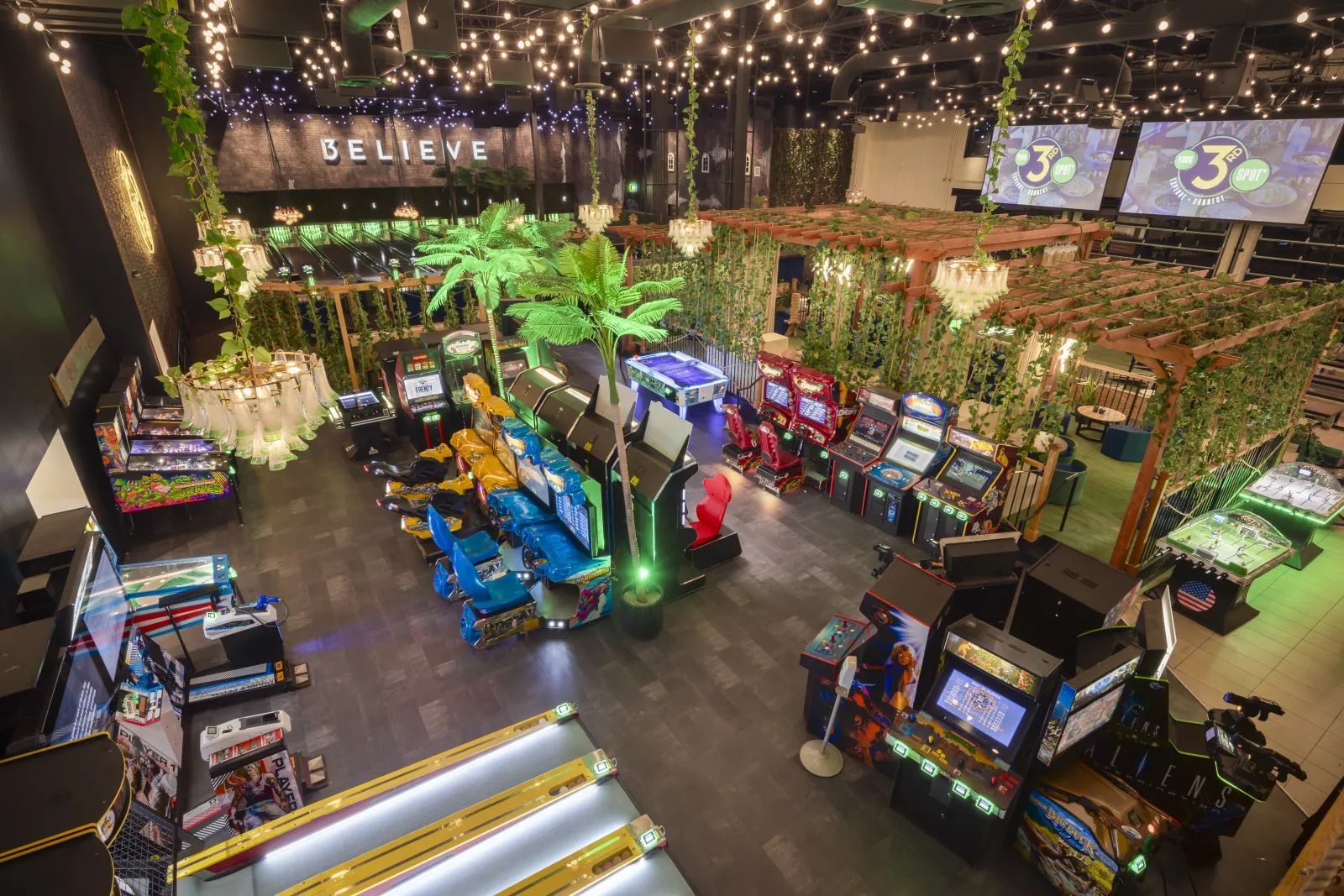 Indoor arcade with vintage and modern game machines, colorful seats, green plants, and string lights above.