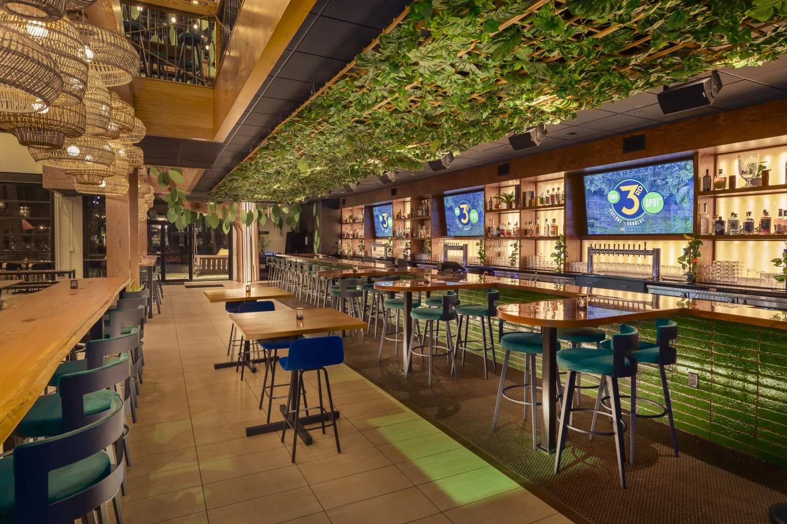 Modern bar interior with green ceiling plants, wooden tables, blue chairs, and illuminated shelves with bottles.