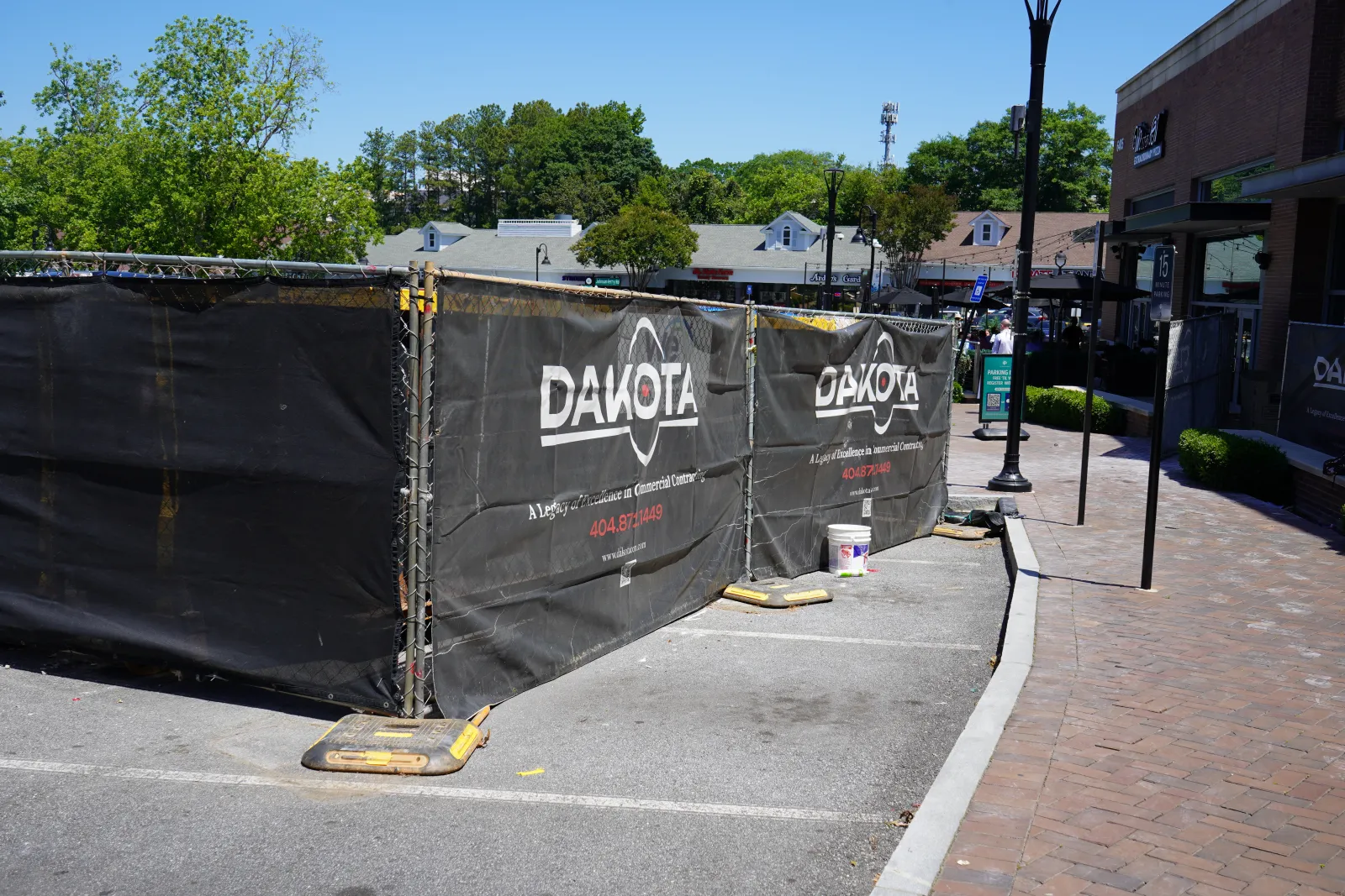 Outdoor construction site fenced with black Dakota-branded mesh screens along a brick sidewalk under clear sky.