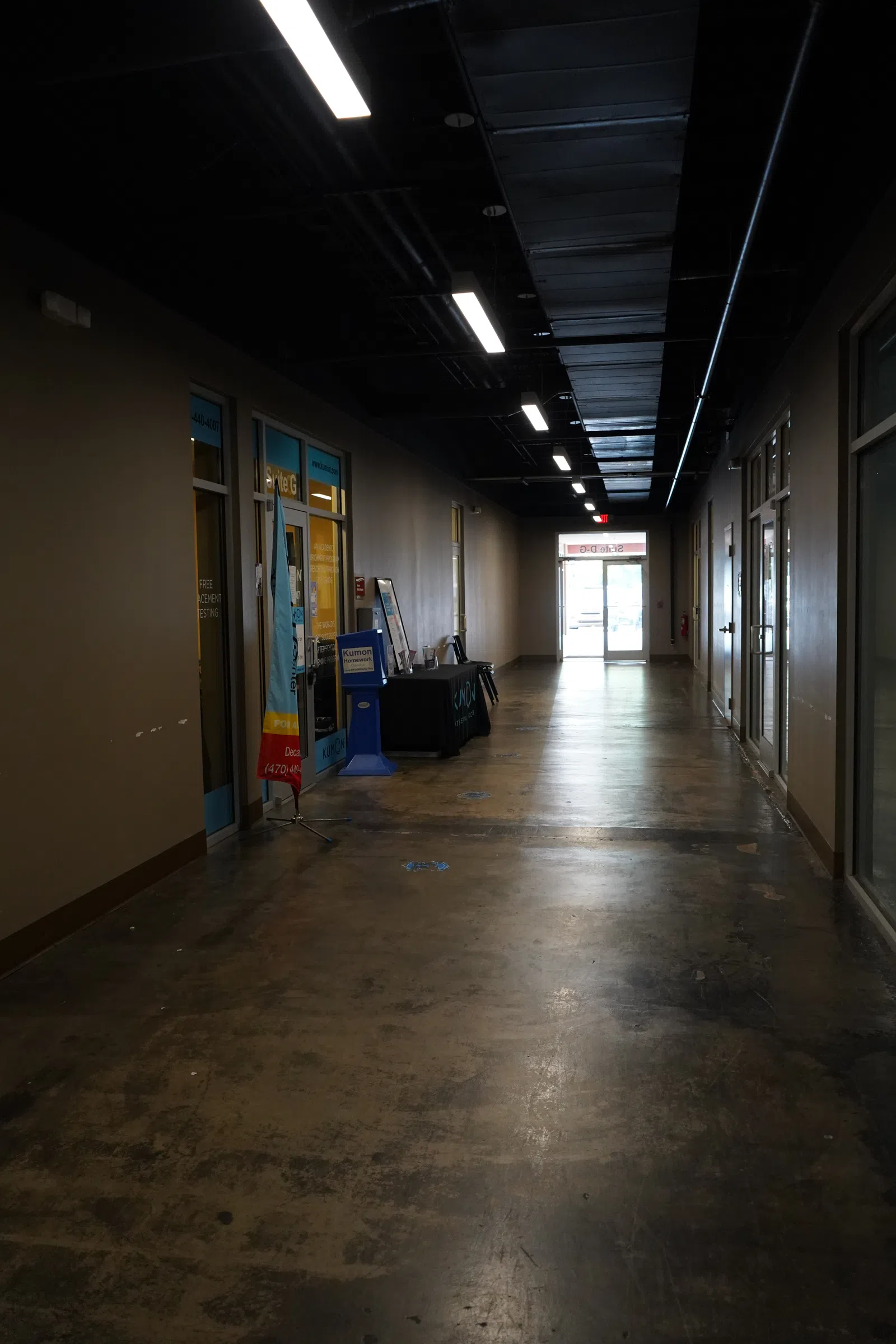Empty commercial hallway with offices, polished concrete floor, overhead lights, and glass doors leading outside.