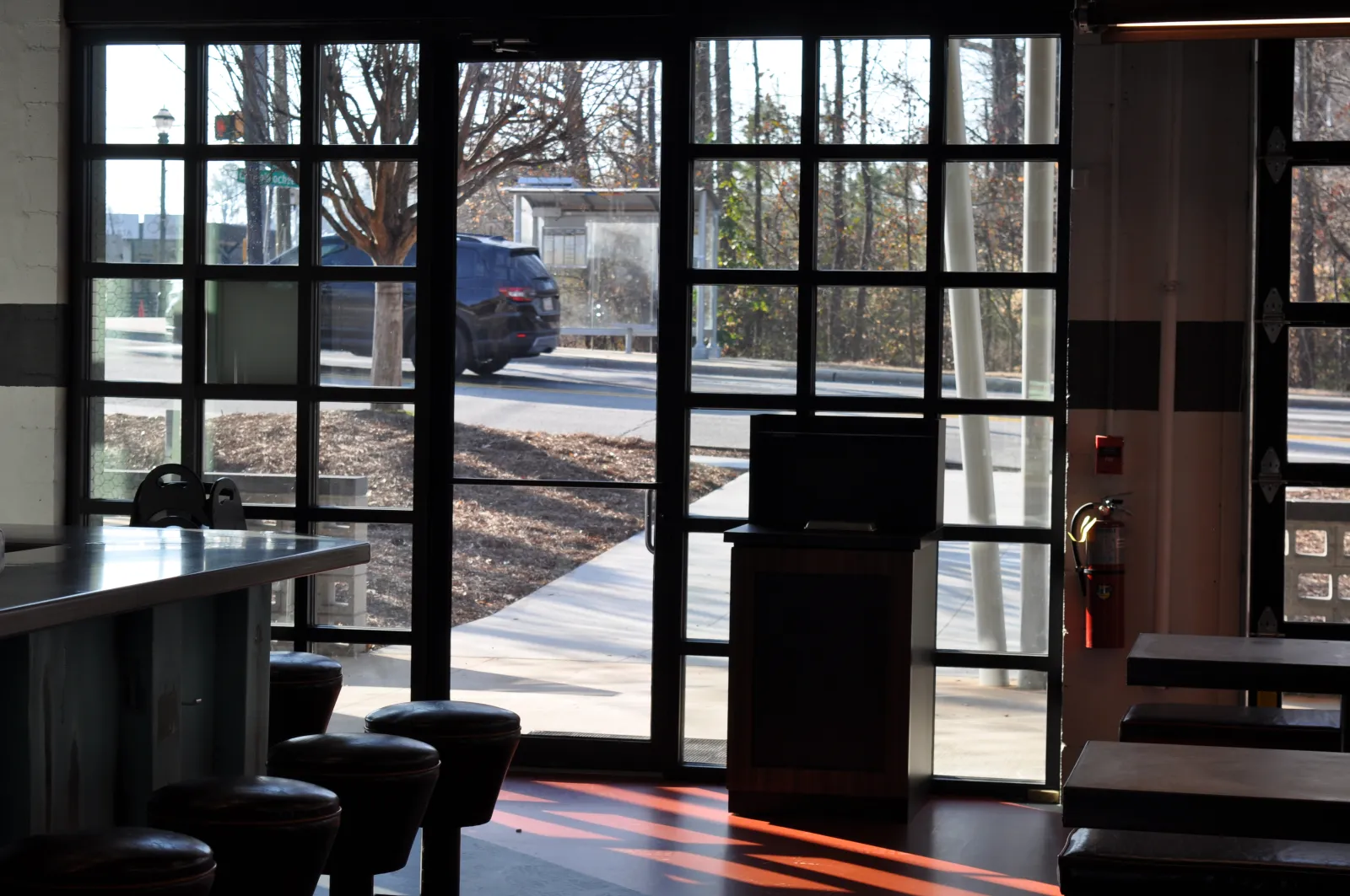 Interior view of a cafe with stools and tables near large glass windows showing an outdoor sidewalk and passing car.