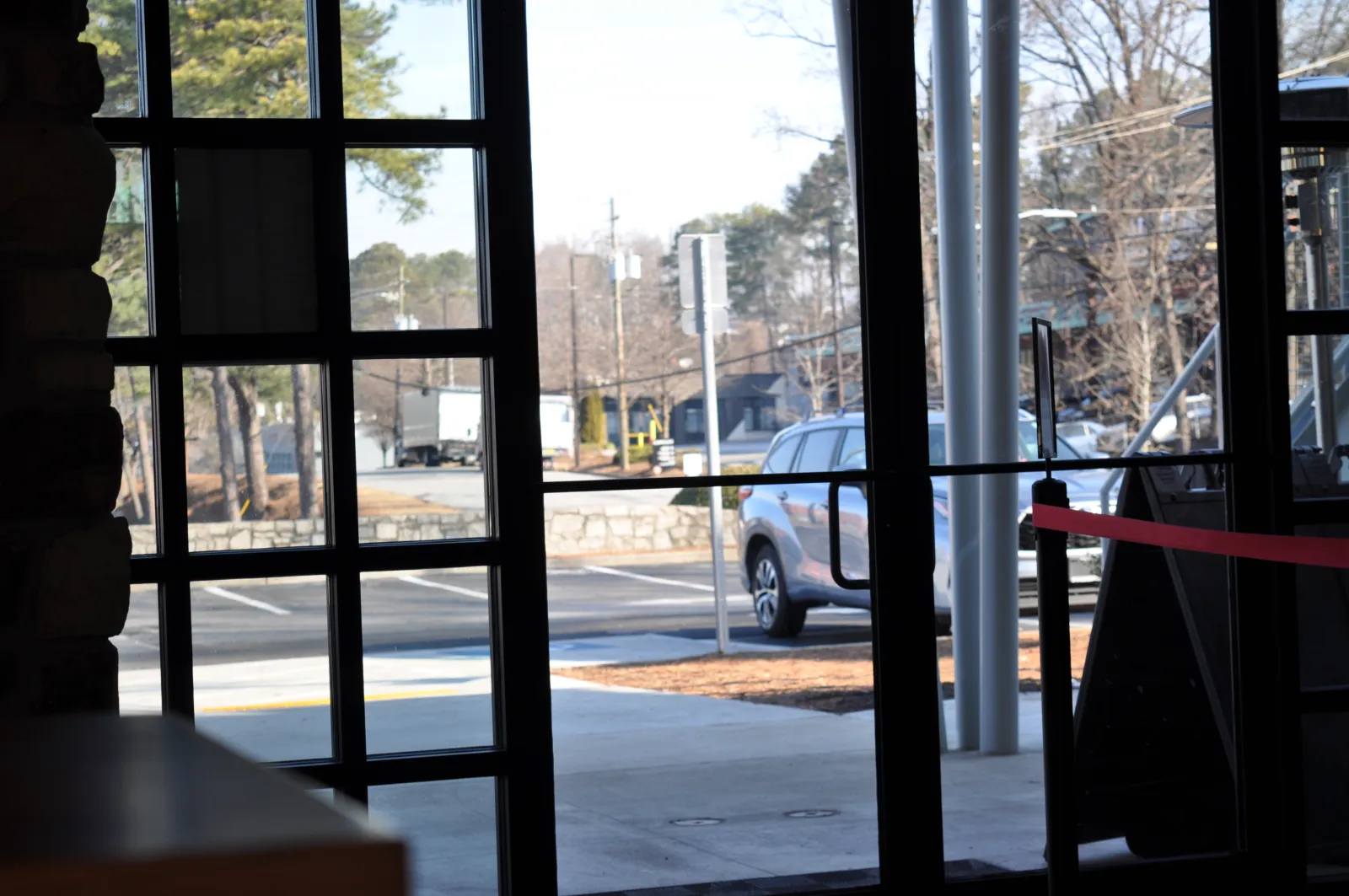 View through glass doors showing a parking lot, a parked silver SUV, and trees on a sunny day.