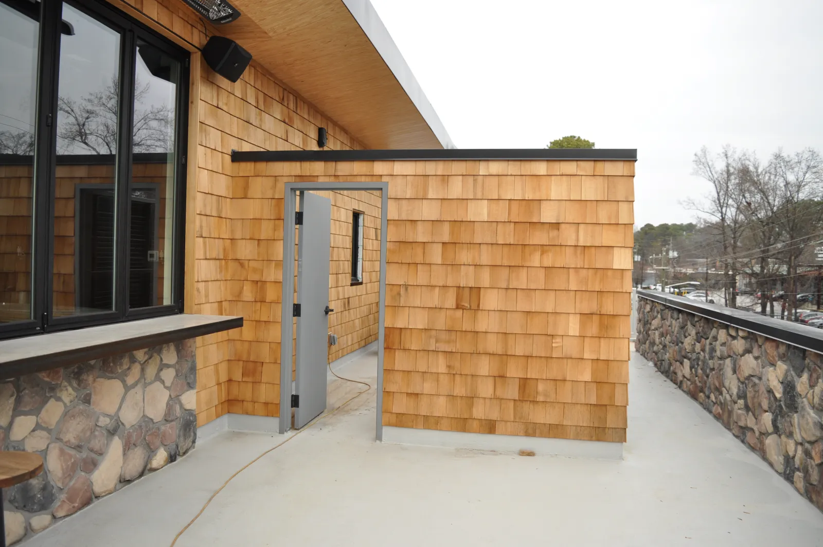Outdoor patio area with wooden shingle walls, stone accents, glass windows, and a metal door leading inside.