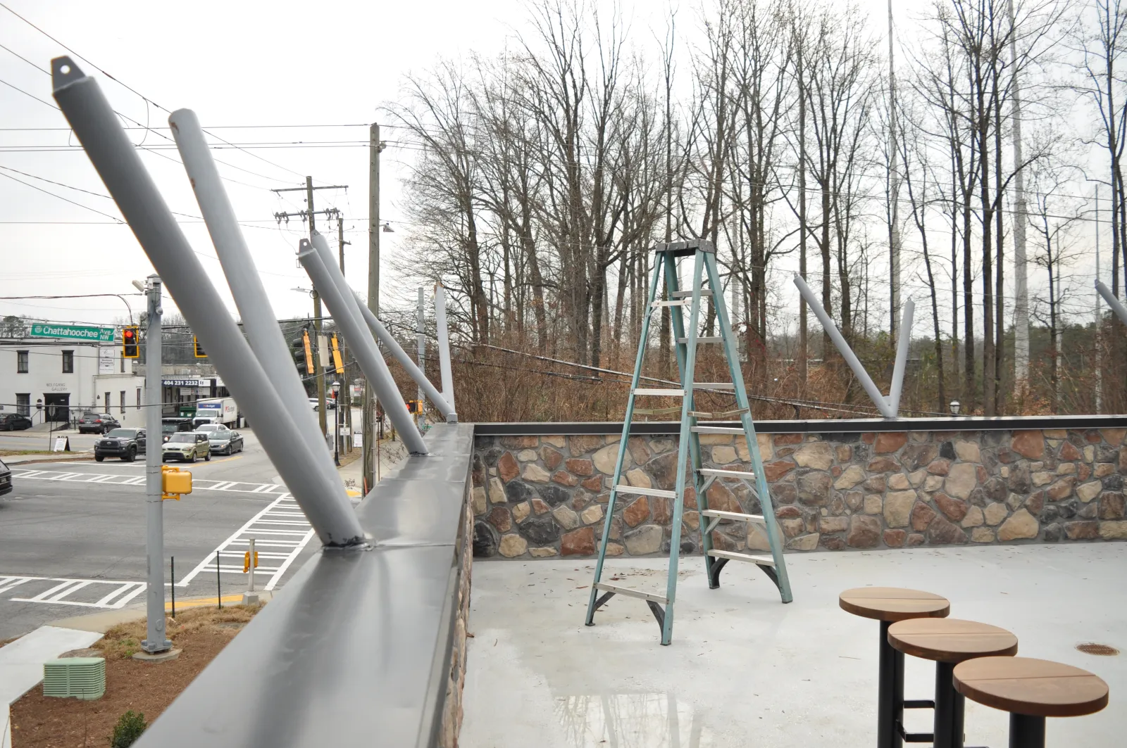 Outdoor patio under construction with metal poles, a green ladder, stone wall, and wood stools on a concrete floor.