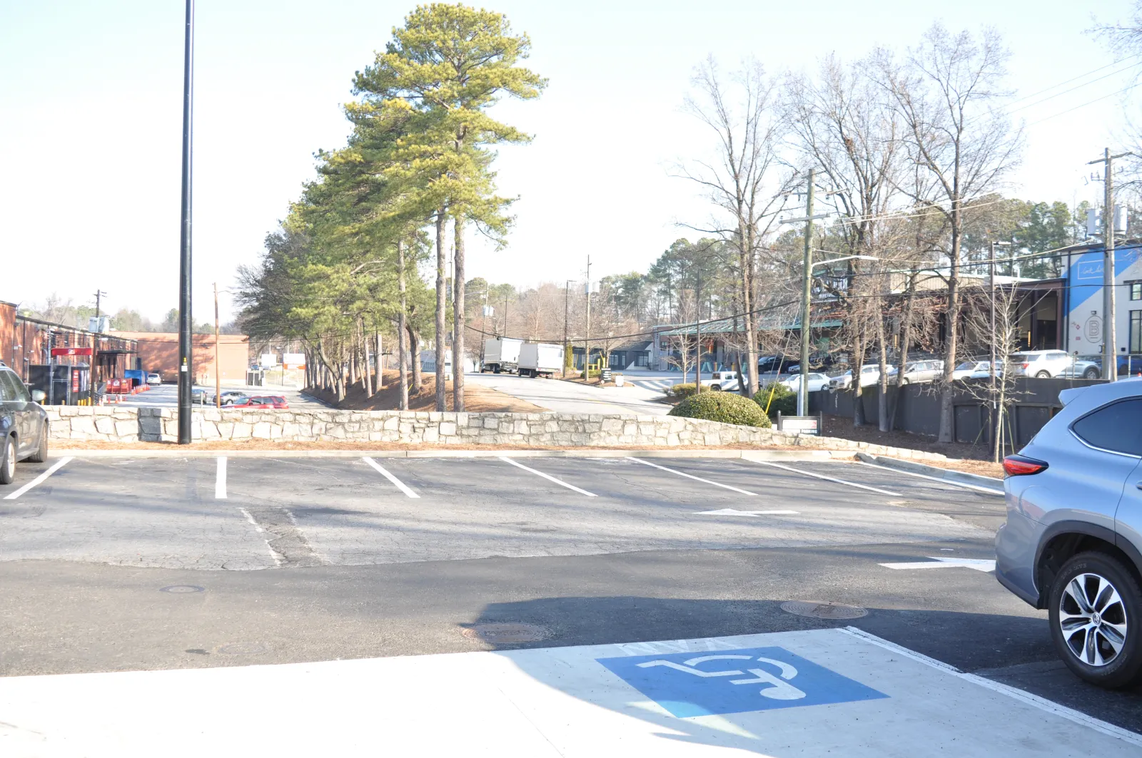 Empty parking lot with a few cars, trees, and buildings in a sunny daytime setting.