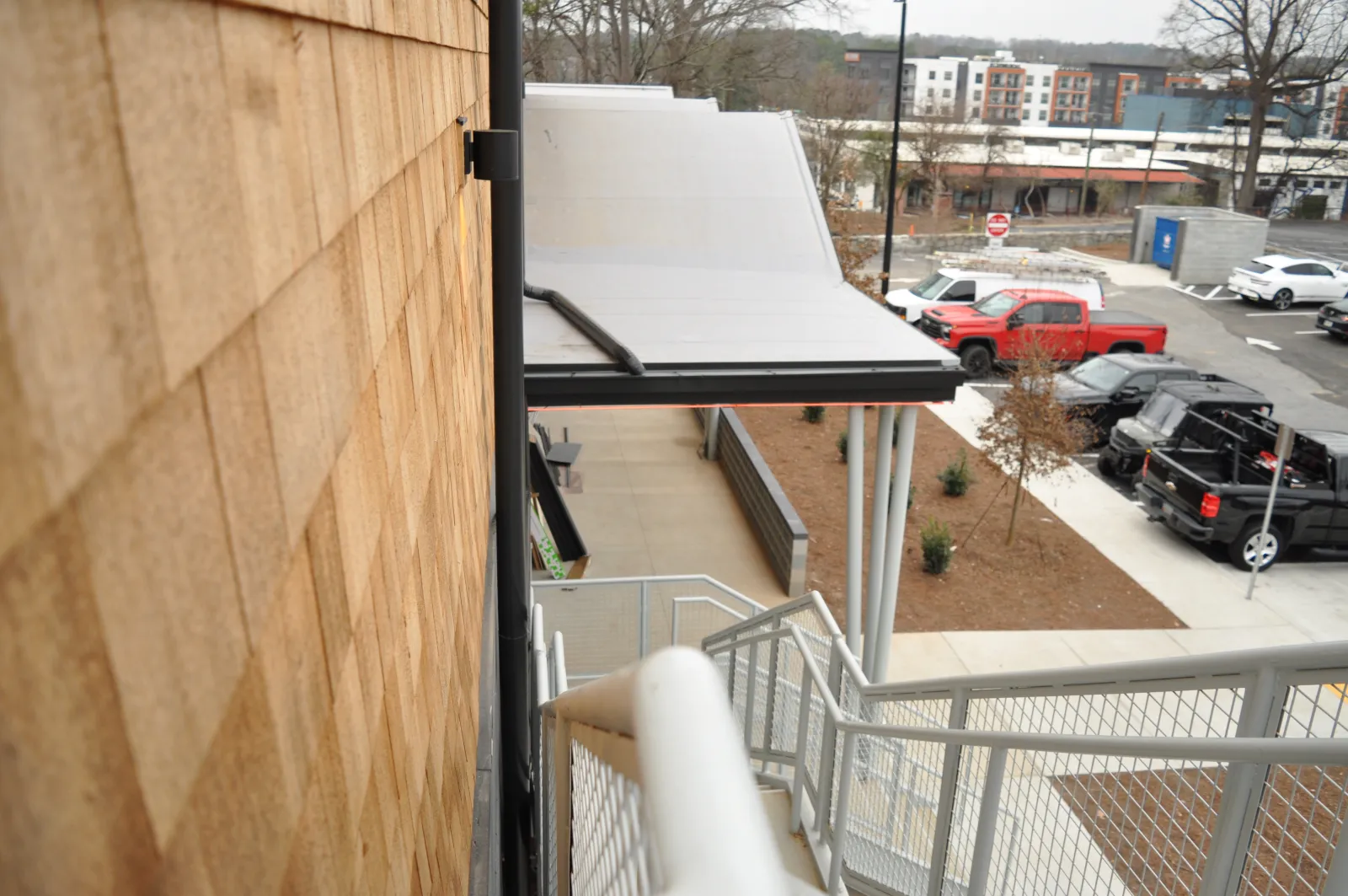 View down a staircase alongside a wooden shingle wall overlooking a parking lot with trucks and buildings in the background.