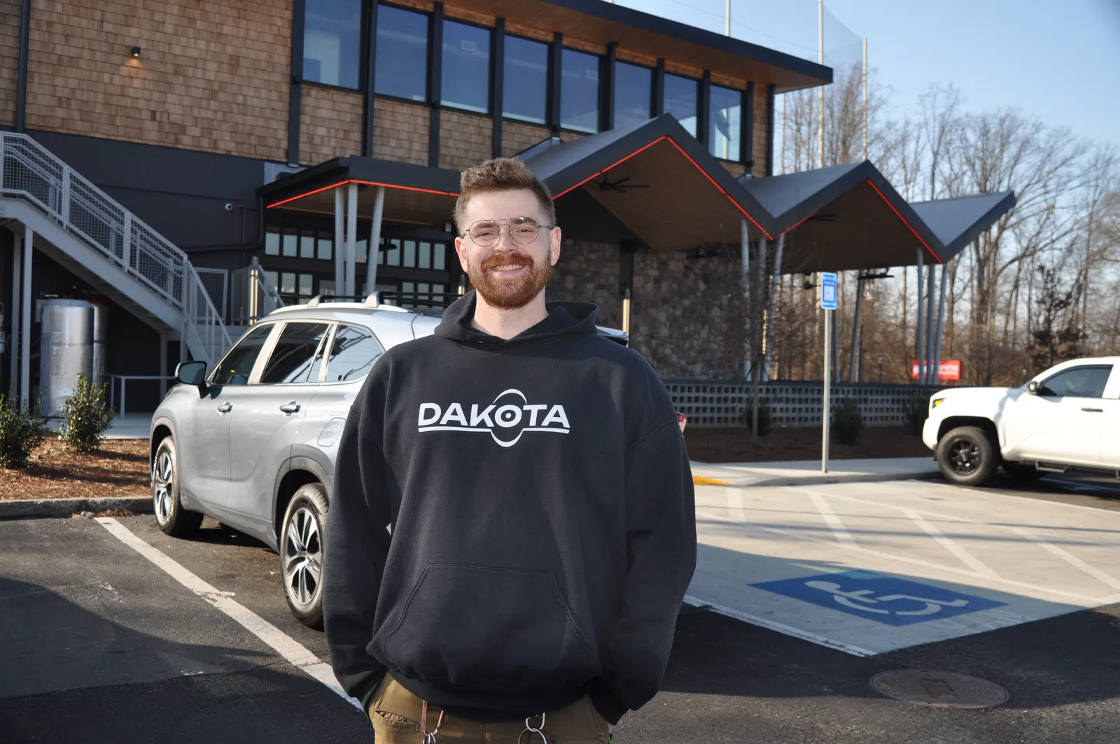 Smiling man in glasses wearing a black Dakota hoodie stands in a parking lot with cars and a modern building in the background.
