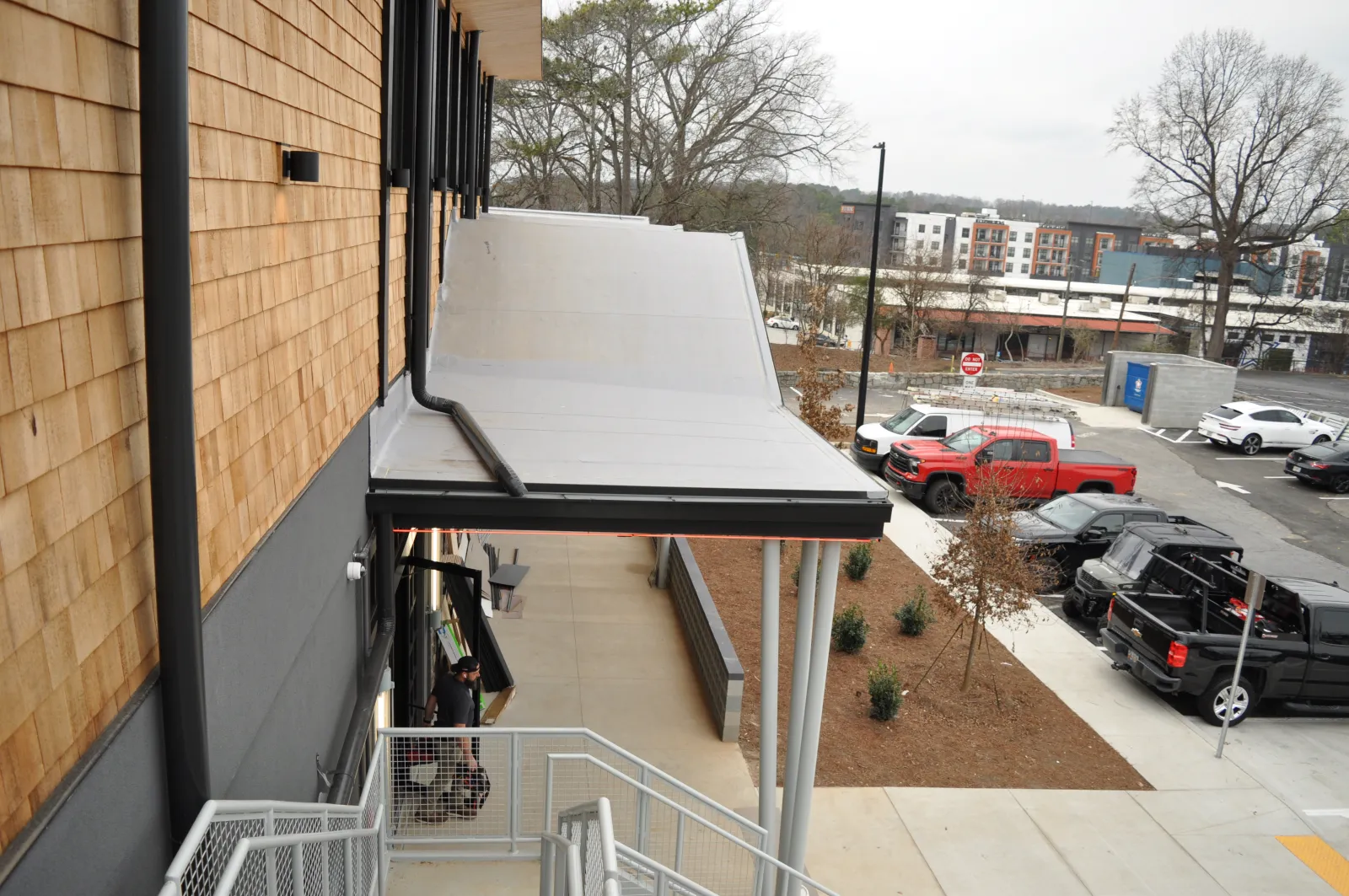 View of modern building exterior with metal awning, staircase, parked trucks, and urban surroundings on a cloudy day