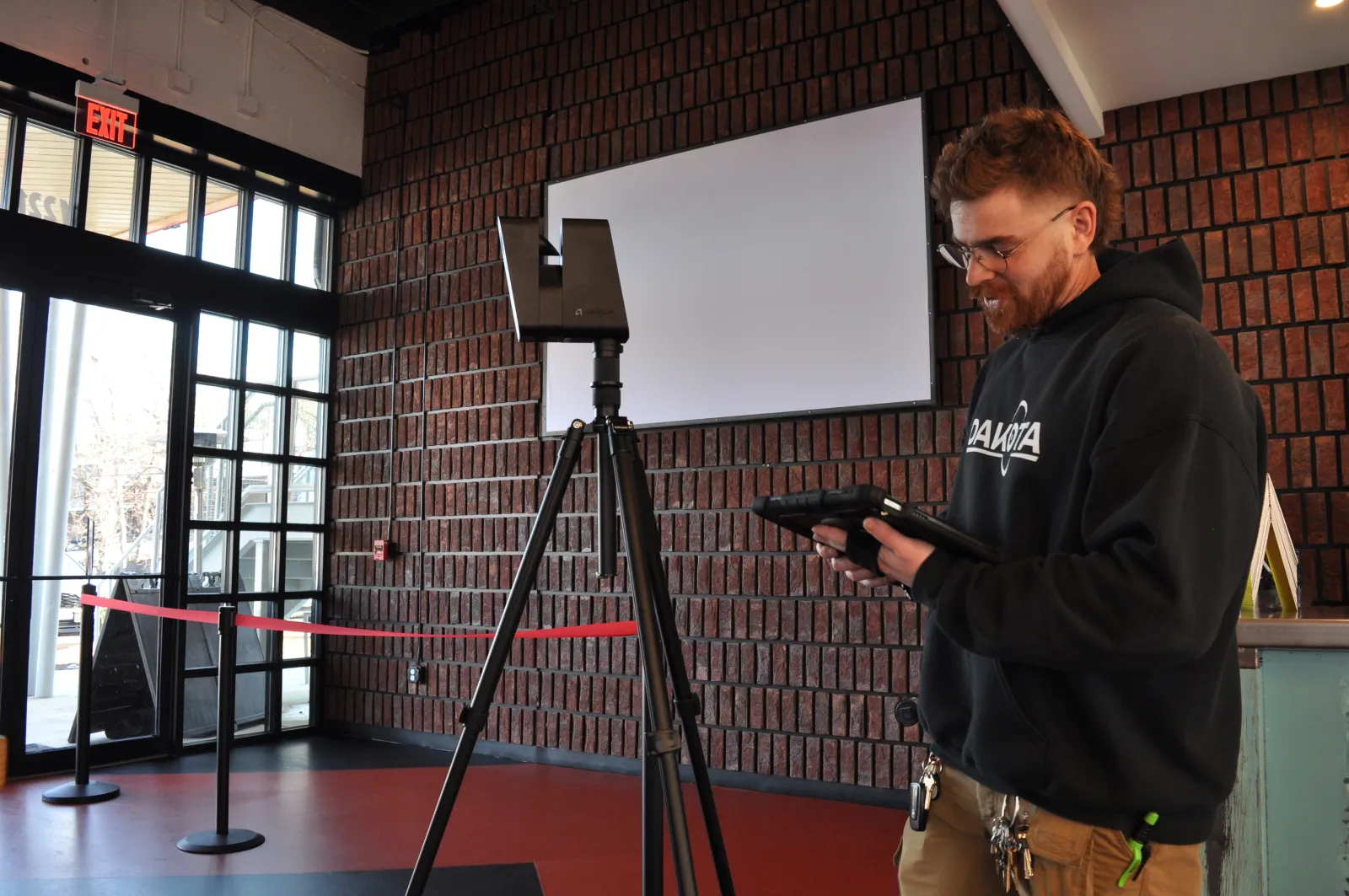 Man operating a 3D scanner on a tripod inside a modern brick-walled room with glass doors and an exit sign.