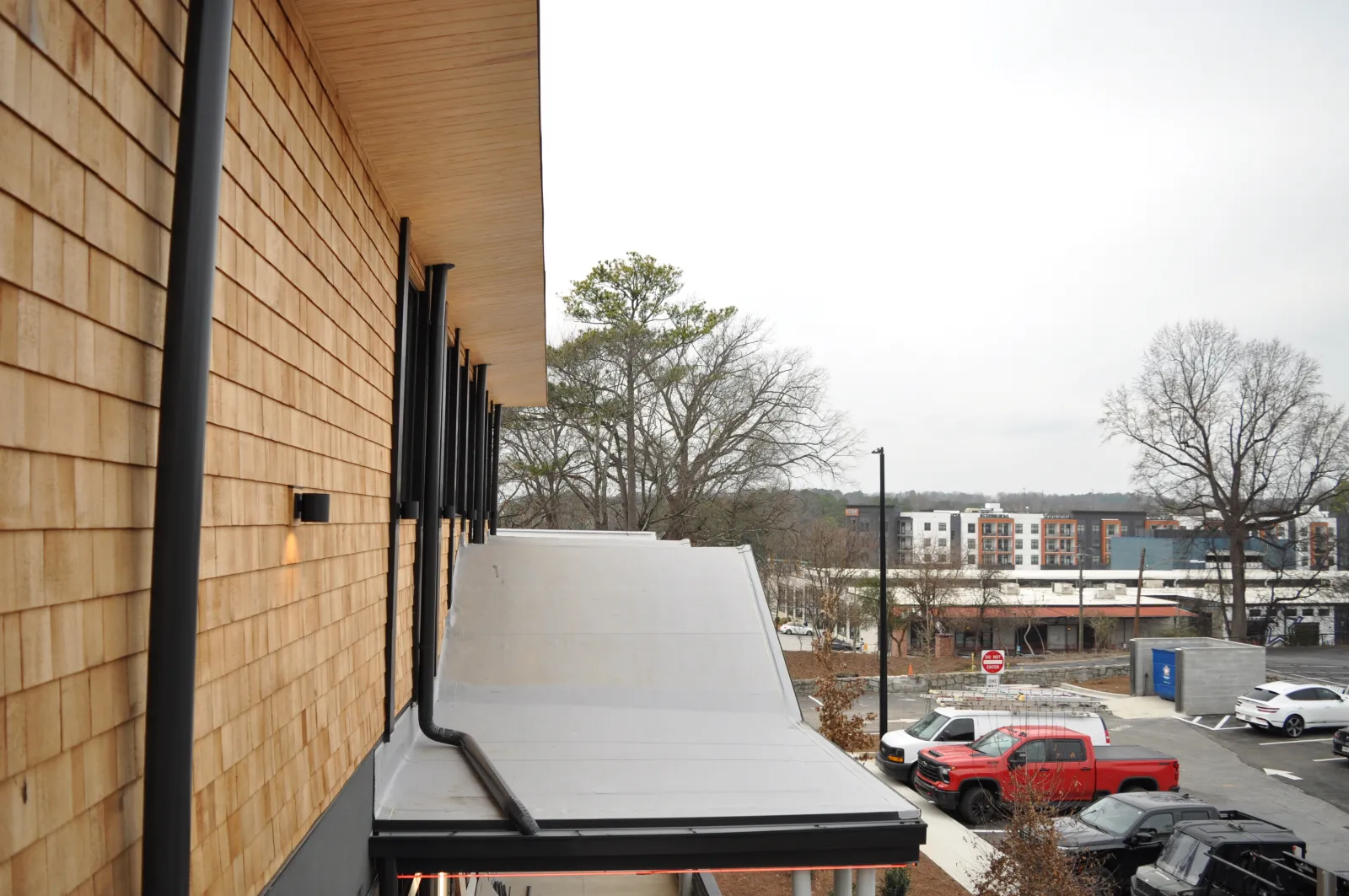 Modern building exterior with wood siding, sloped gray metal awnings, and a parking lot with vehicles and trees in the background