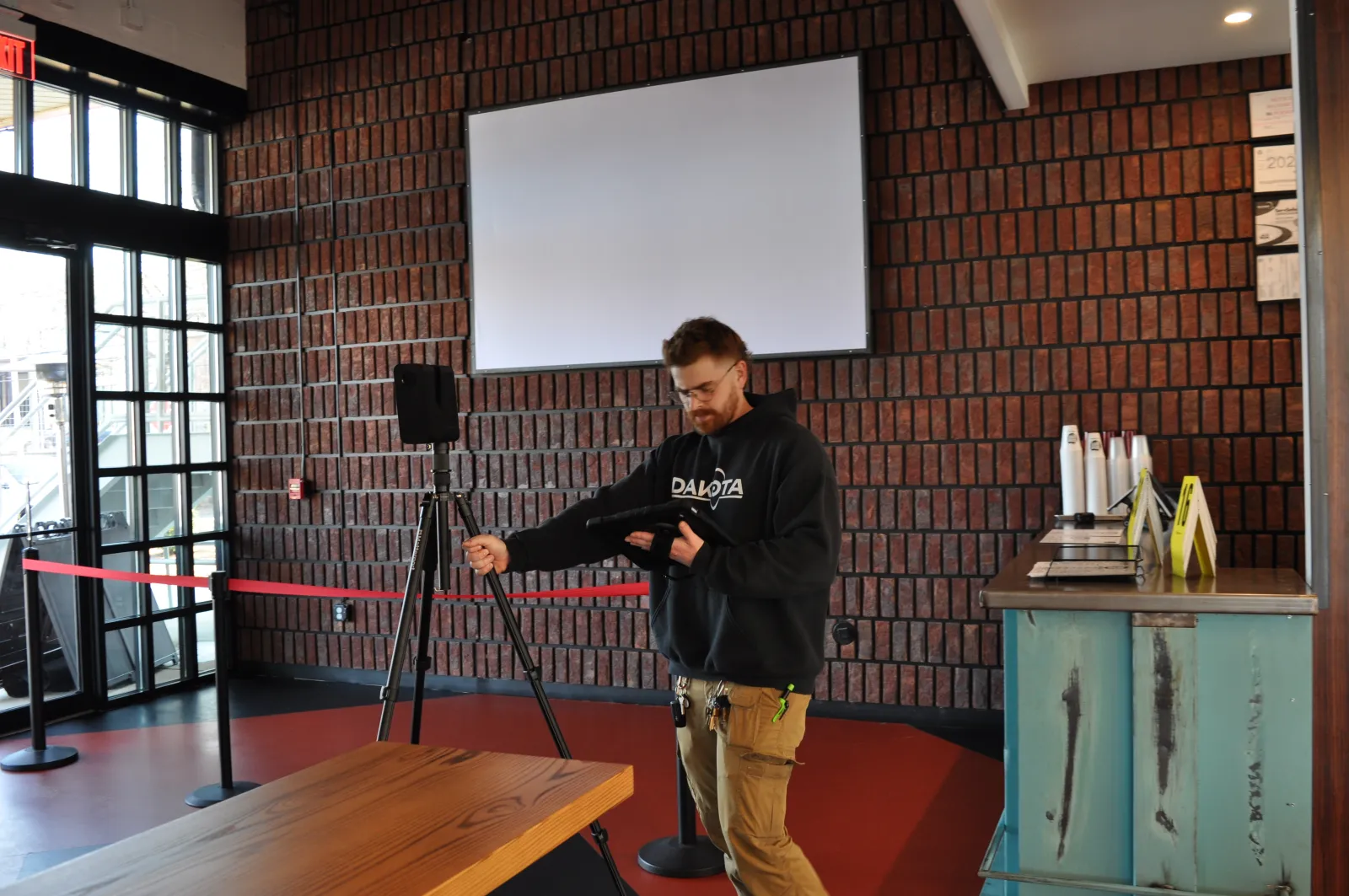 Man adjusting a tripod-mounted device in a modern room with brick walls and large windows.