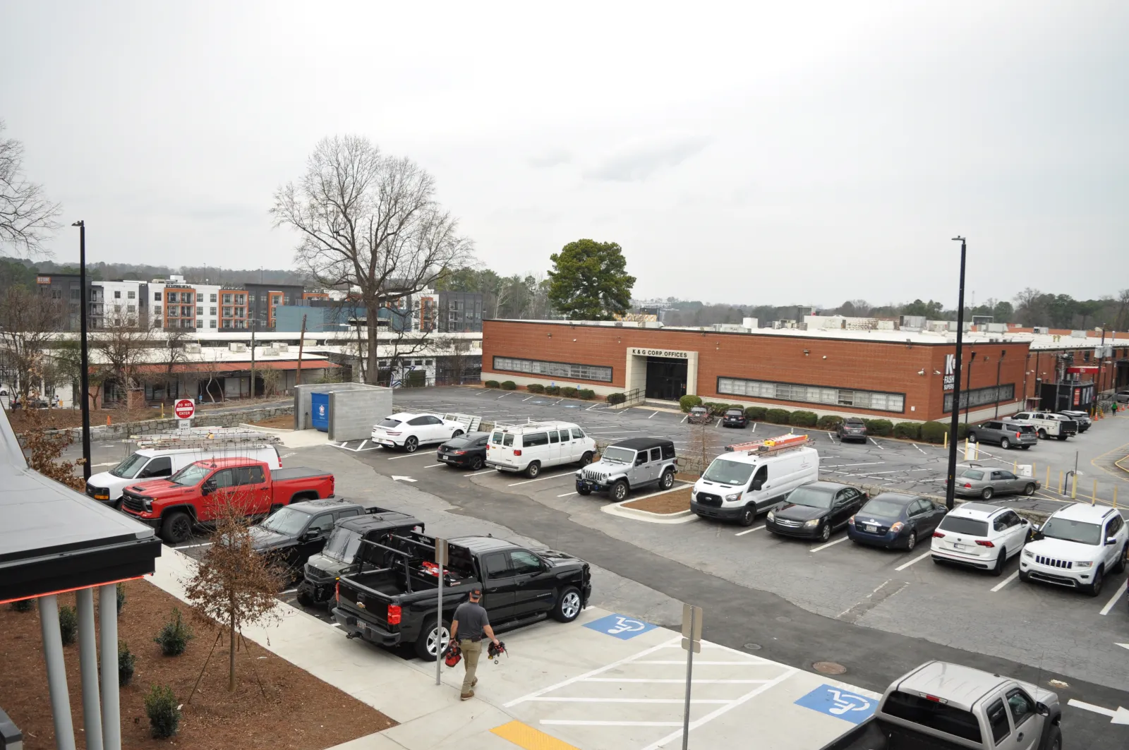 Urban parking lot with various vehicles, including trucks and vans, in front of a brick commercial building on a cloudy day.