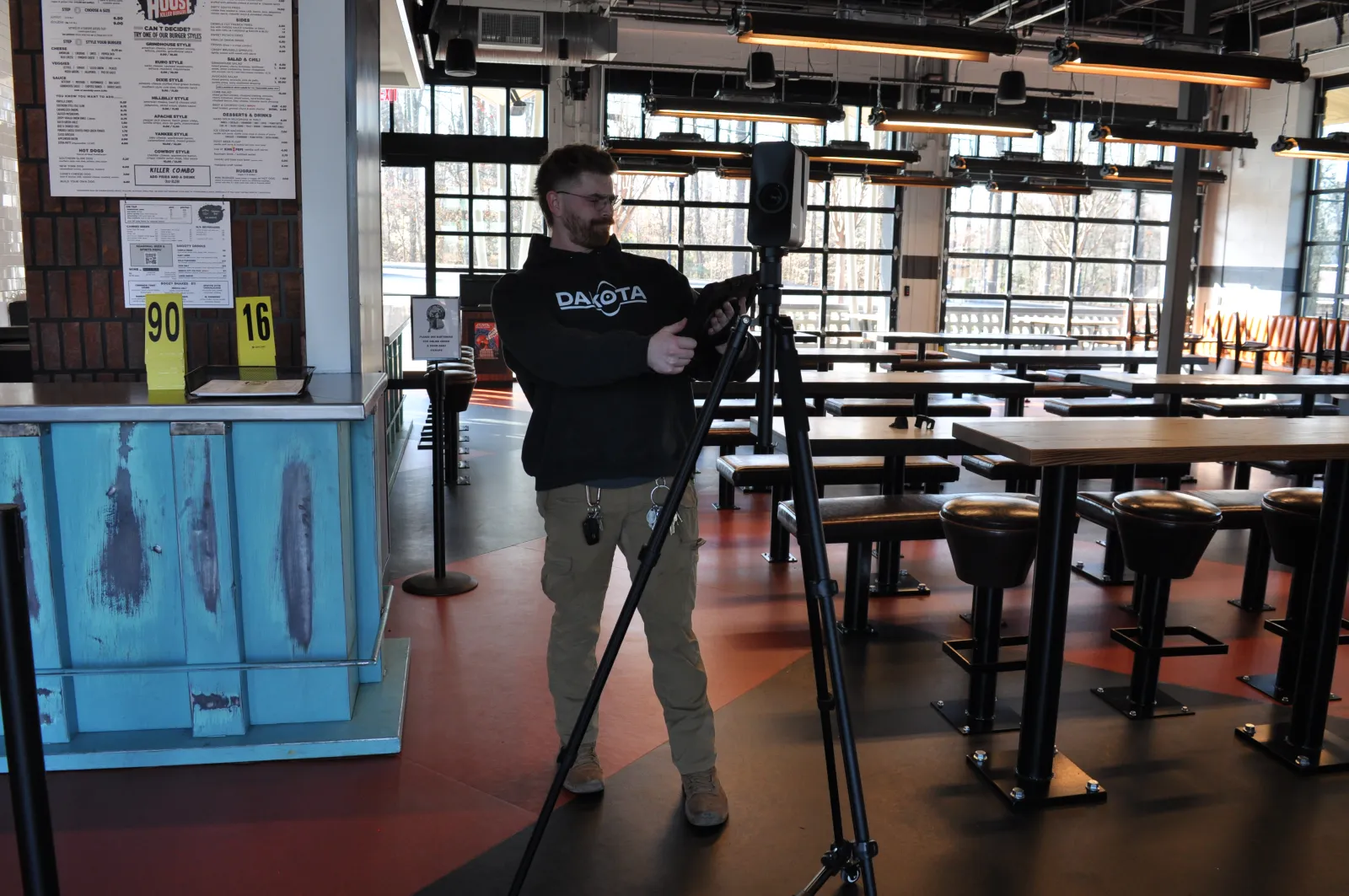 Man adjusting camera on tripod inside a modern cafe with empty tables and bar stools during daytime.