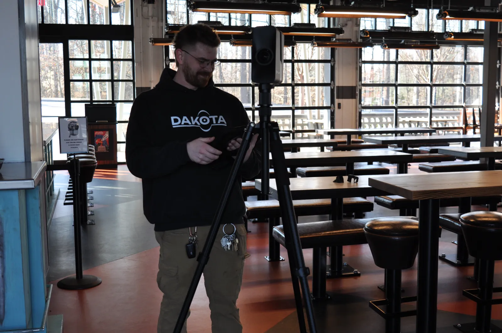 Man adjusting camera equipment on tripod inside a modern empty restaurant with large windows and tables.