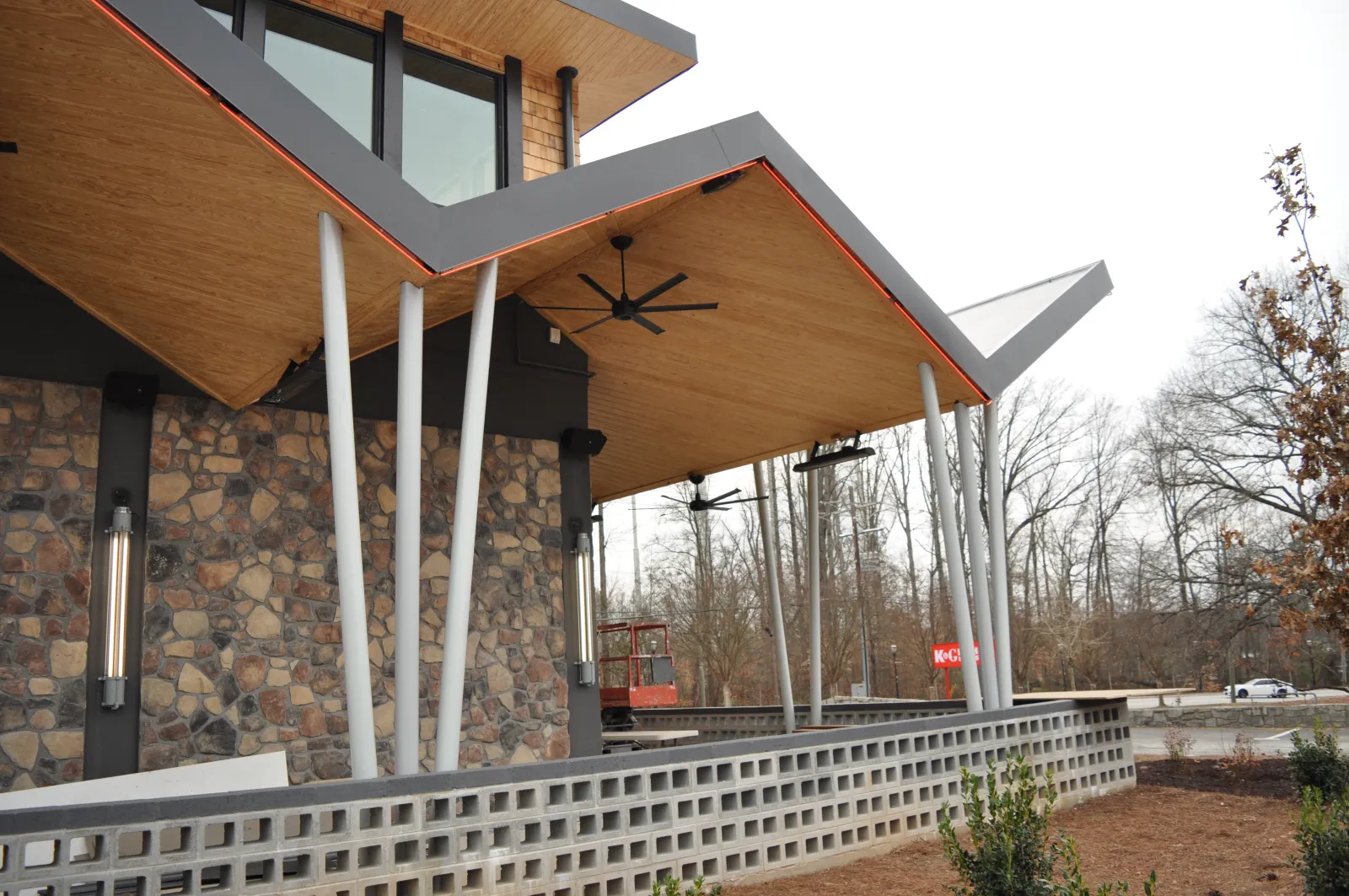 Modern building exterior with angled wooden roof, white pillars, stone wall, and outdoor ceiling fans overlooking a garden.