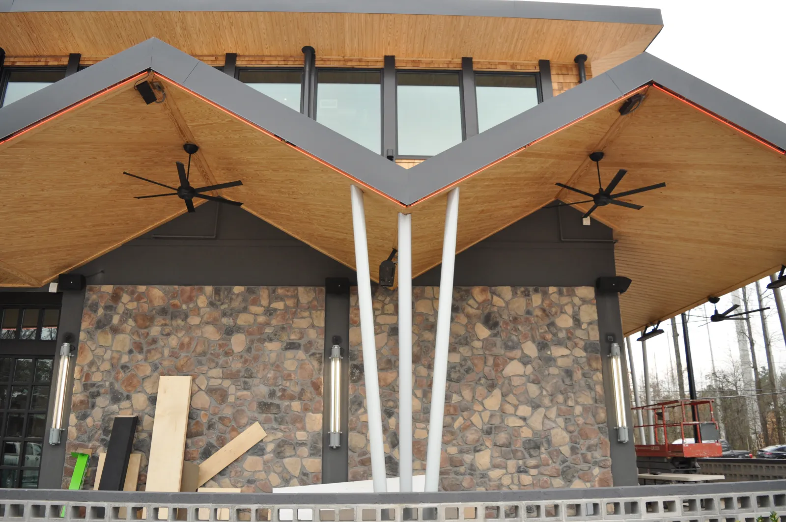 Modern building entrance with unique angular wooden roof, stone wall, ceiling fans, and metal support columns.