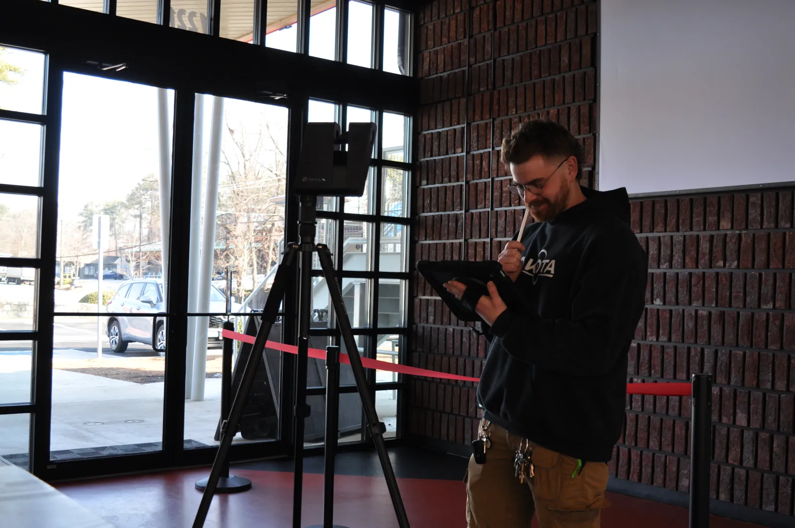 Man wearing glasses working on a tablet device next to tripod-mounted equipment inside a building lobby.