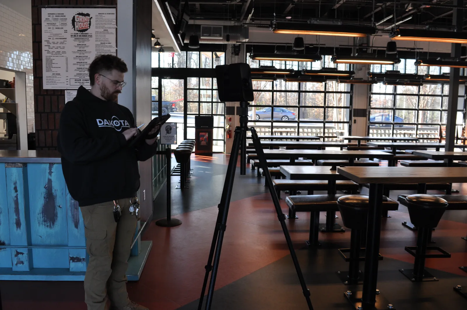 Man wearing Dakota sweatshirt using tablet near a tripod in a spacious, modern restaurant with large windows.