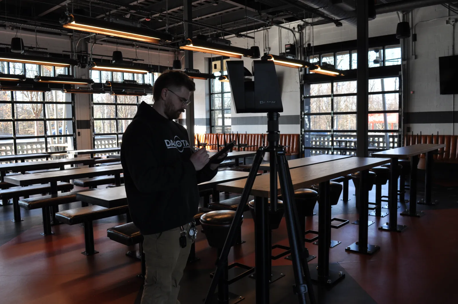 Man in a black hoodie using a tablet next to a speaker on a tripod inside a modern empty restaurant with wooden tables.