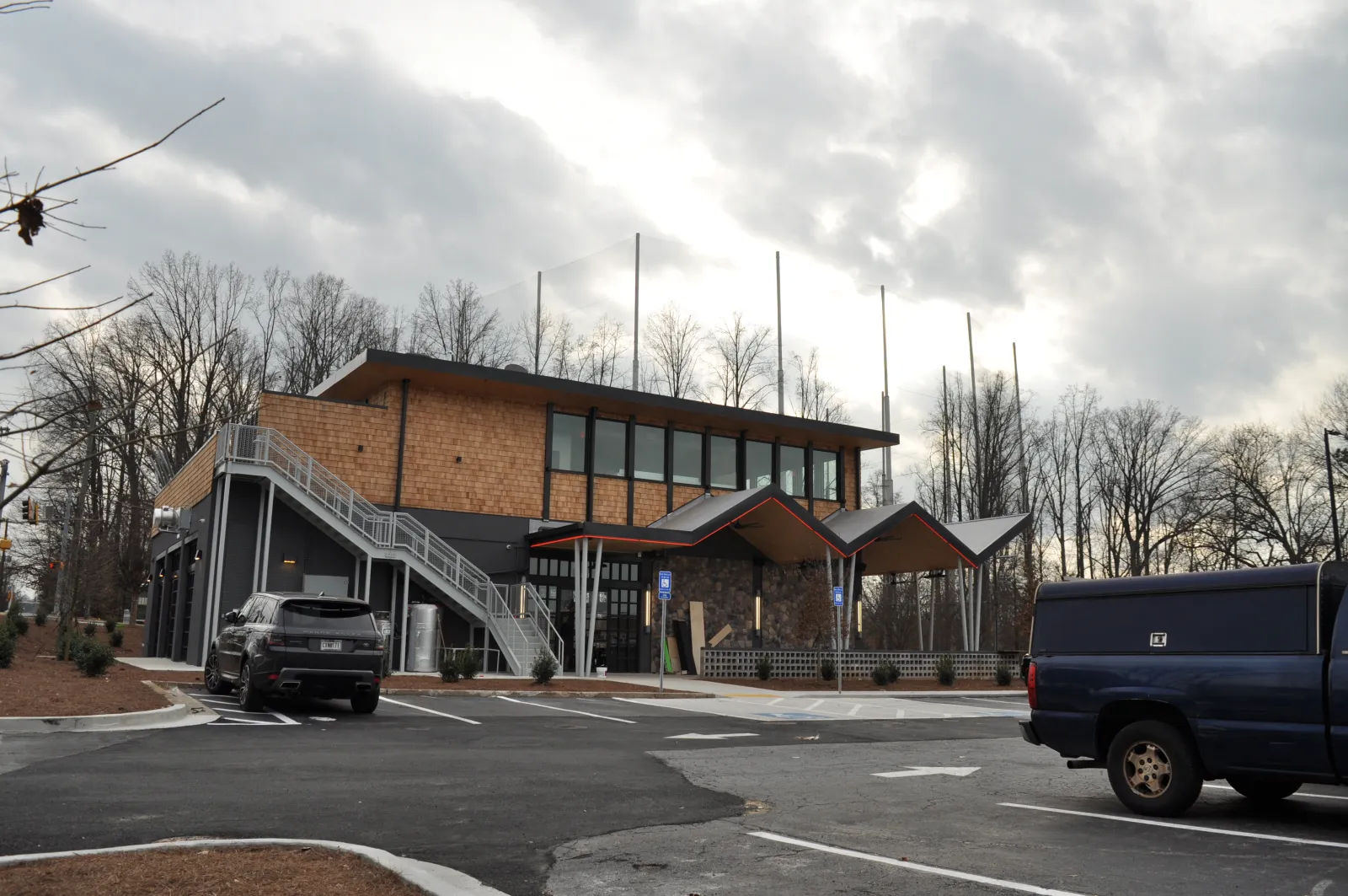 Modern two-story building with large windows and outdoor staircase in a parking lot under cloudy sky.