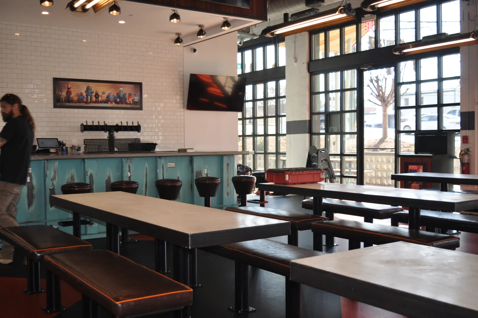 Modern indoor bar with long tables, leather benches, bar stools, large windows, and industrial lighting fixtures.