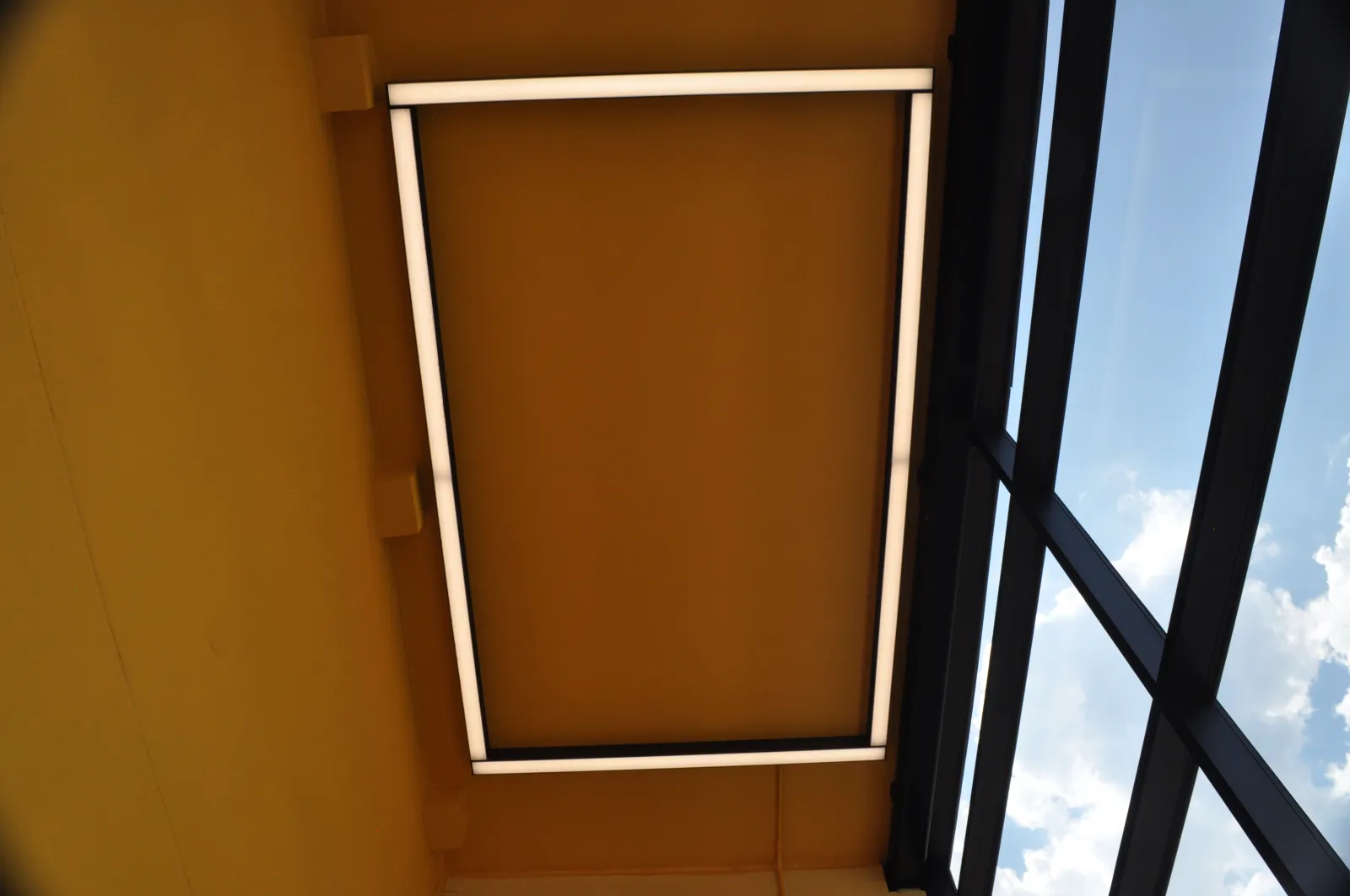Modern rectangular ceiling light fixture glowing against an orange ceiling next to a glass window with blue sky.