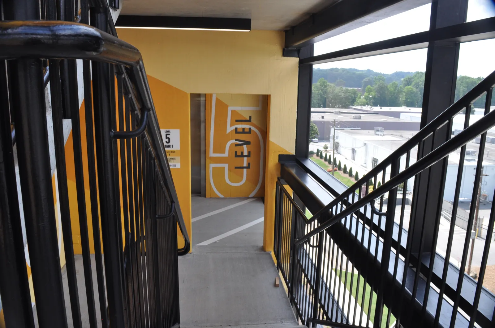 Staircase leading to level 5 of a parking garage with yellow walls and large windows overlooking an industrial area.