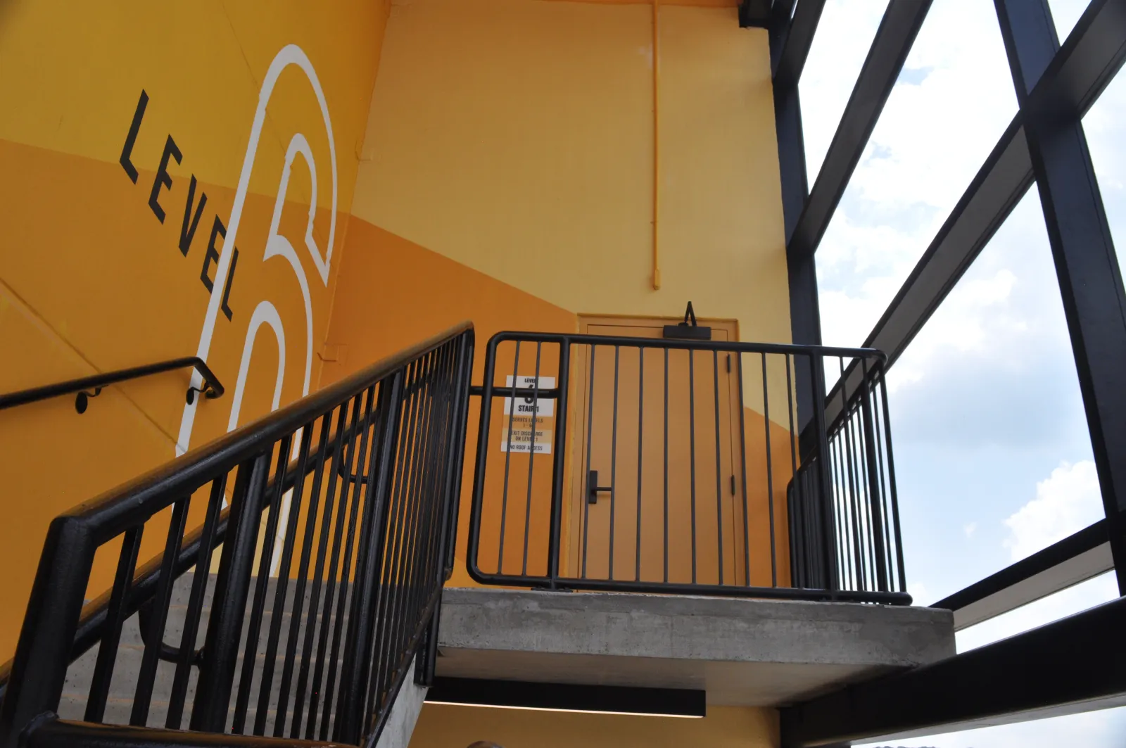 Stairwell leading to Level 6 with yellow walls, black railings, and large windows showing blue sky and clouds.