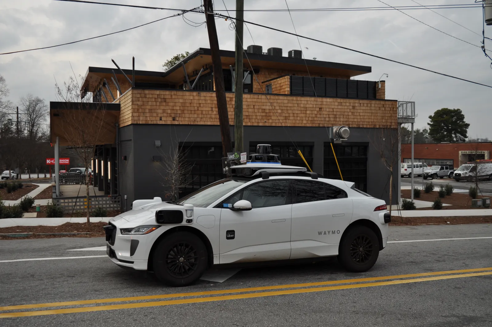 White Waymo self-driving electric car parked on a city street with modern building in the background.