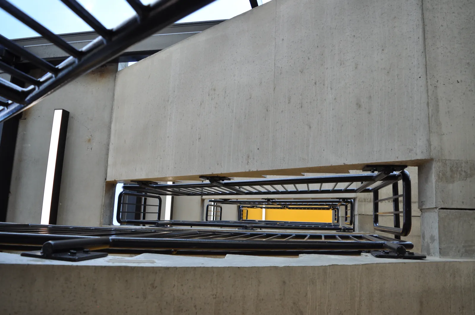 View looking down a modern concrete stairwell with black metal railings and a lit vertical light fixture.