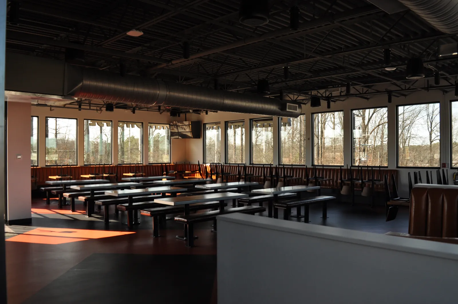 Empty modern cafeteria with long tables, benches, large windows, and exposed ceiling pipes in natural light.