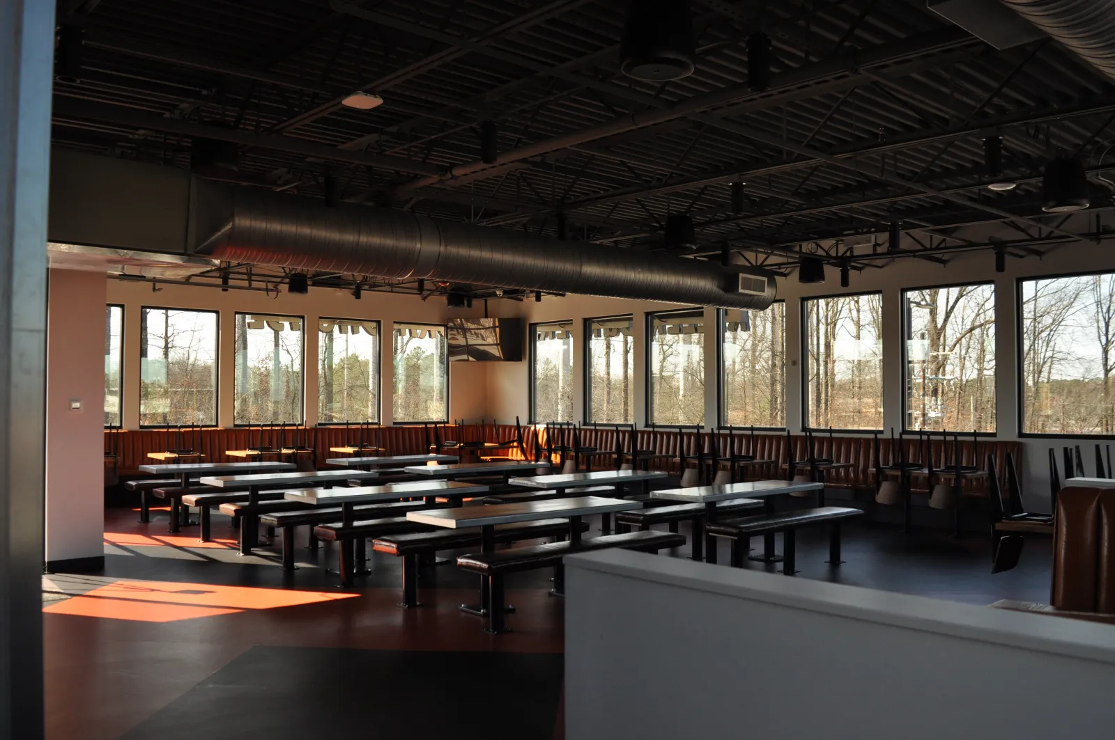 Empty modern cafeteria with long tables, benches, large windows, and exposed ceiling ducts in daylight.