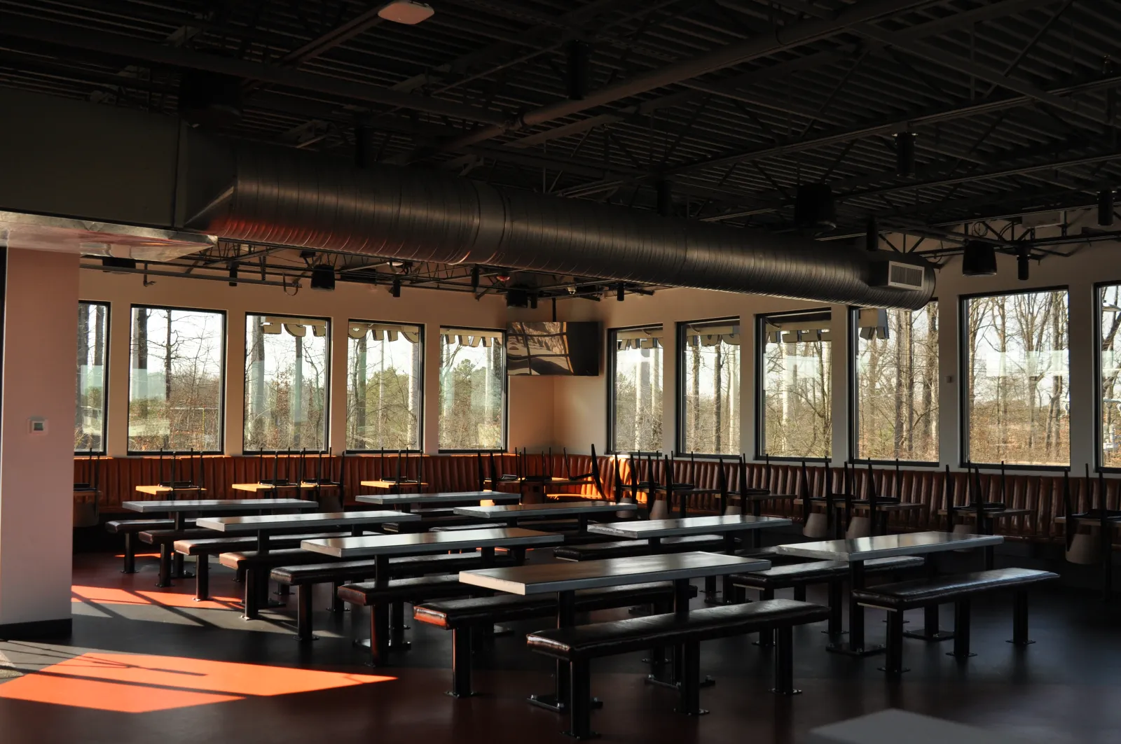 Empty modern cafeteria with large windows, wooden benches, and overhead ventilation ducts during daytime.