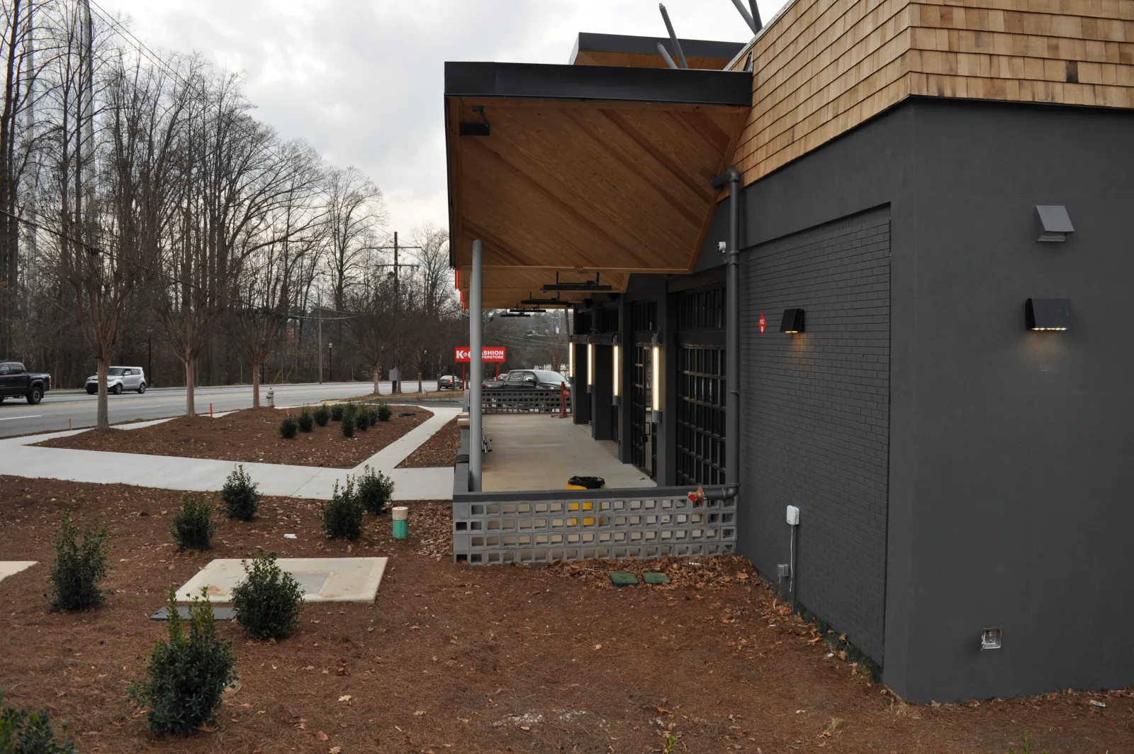Modern building exterior with wooden and black brick facade beside a bare landscaped area and road with vehicles.