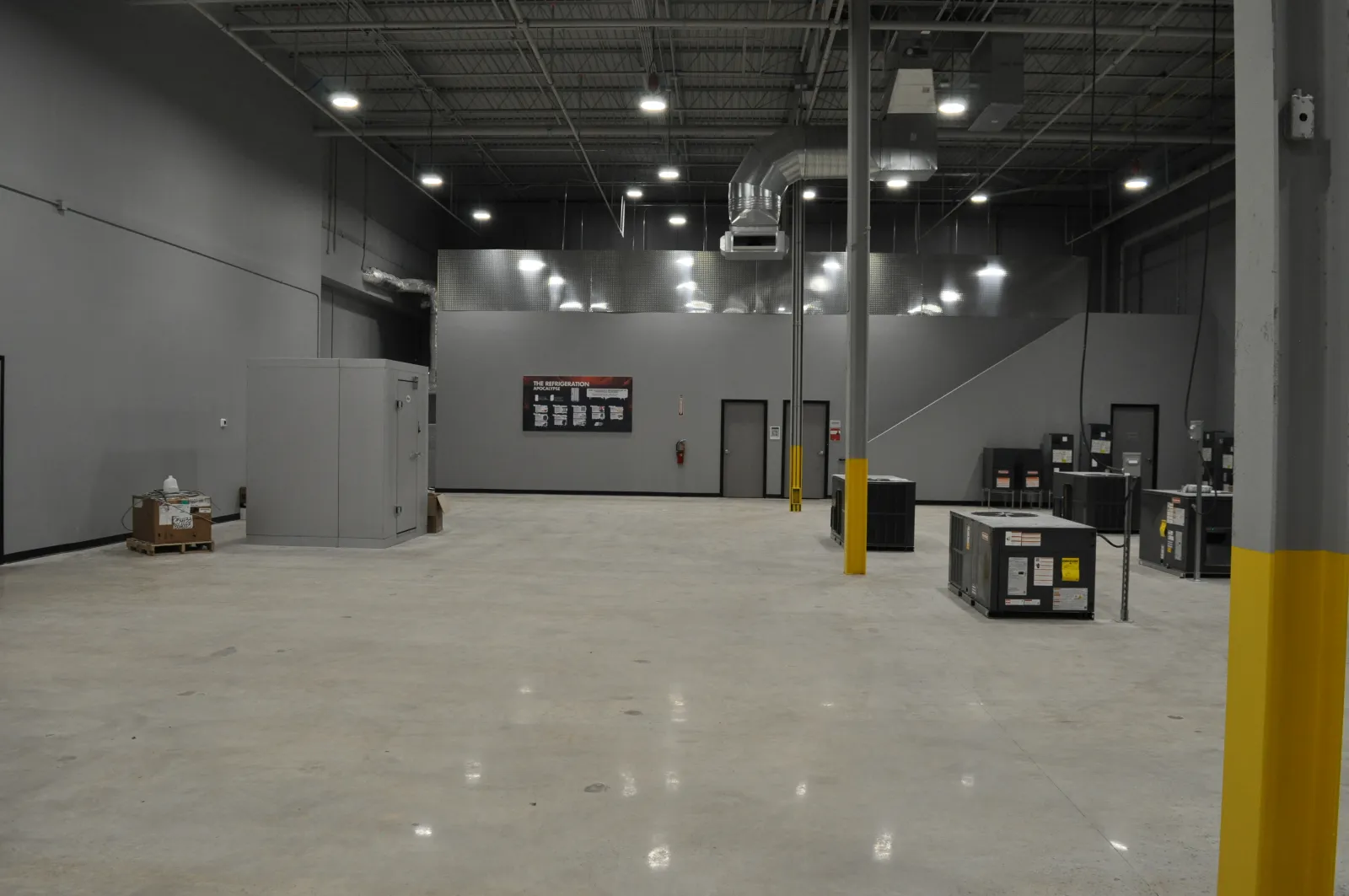 Spacious industrial warehouse interior with gray walls, concrete floor, HVAC units, and bright overhead lights.