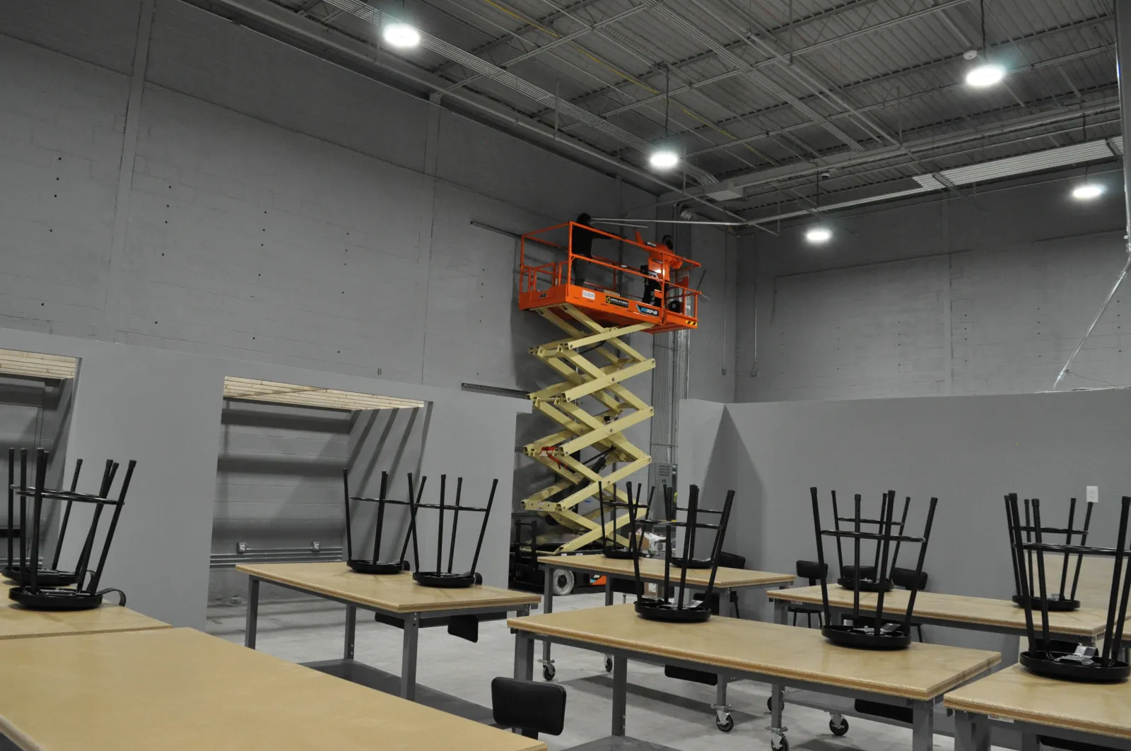 Scissor lift elevating two workers inside a large industrial room with tables and chairs stacked upside down.