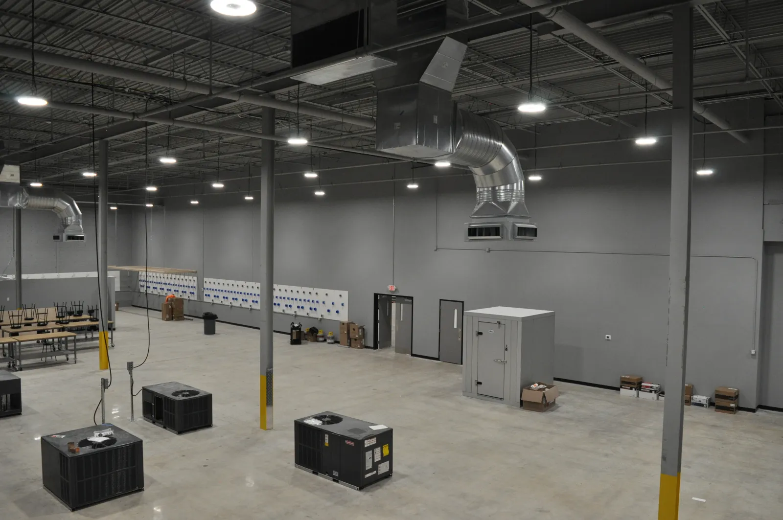 Spacious empty industrial warehouse with concrete floor, large HVAC units, and gray walls under bright ceiling lights.
