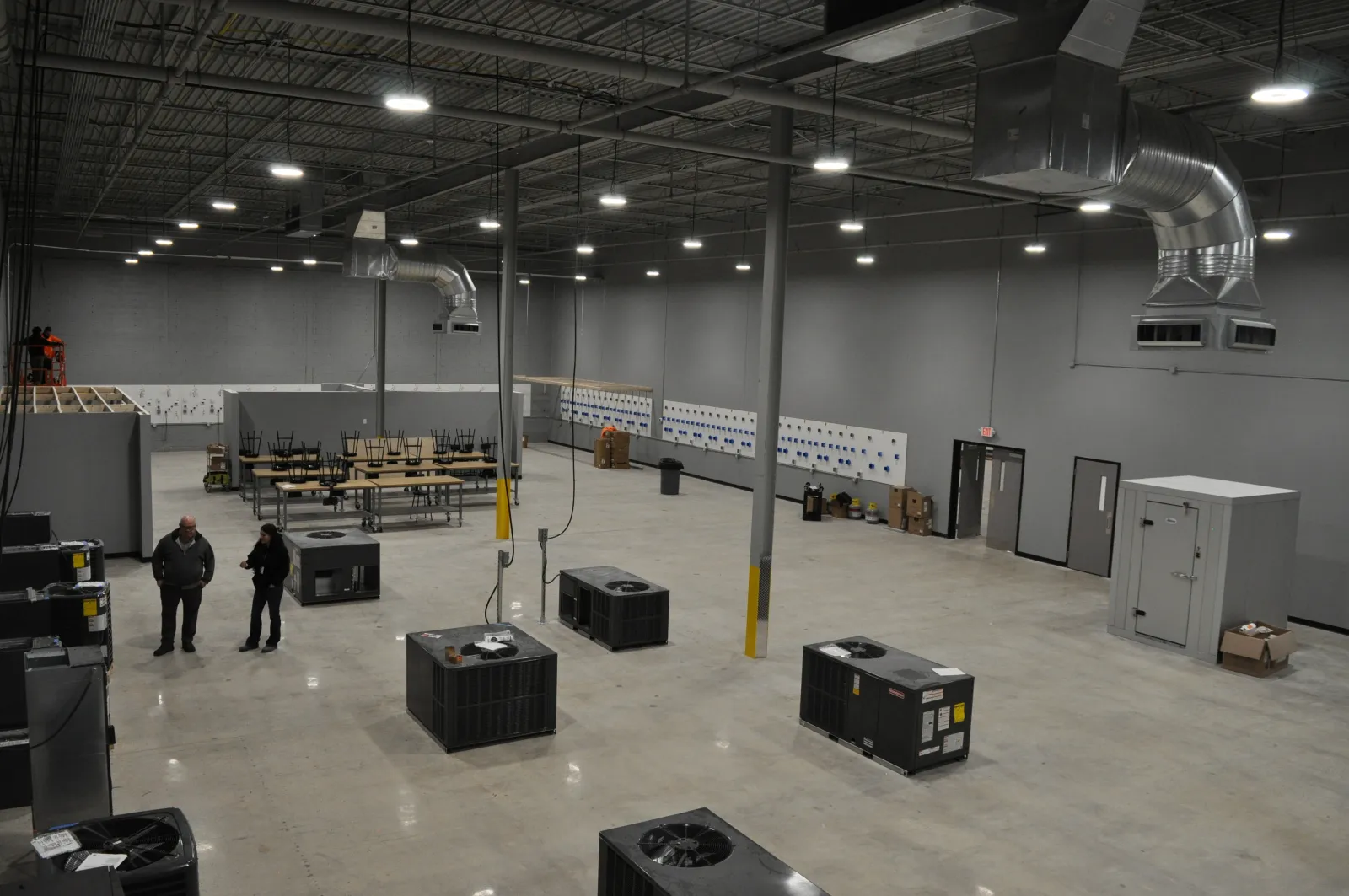 Spacious industrial warehouse with HVAC units on floor, high ceilings, ventilation ducts, two people and tables in background