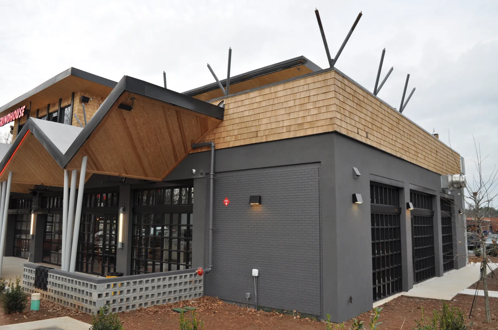 Modern restaurant exterior with gray brick, large windows, and angled wooden roof under cloudy sky.