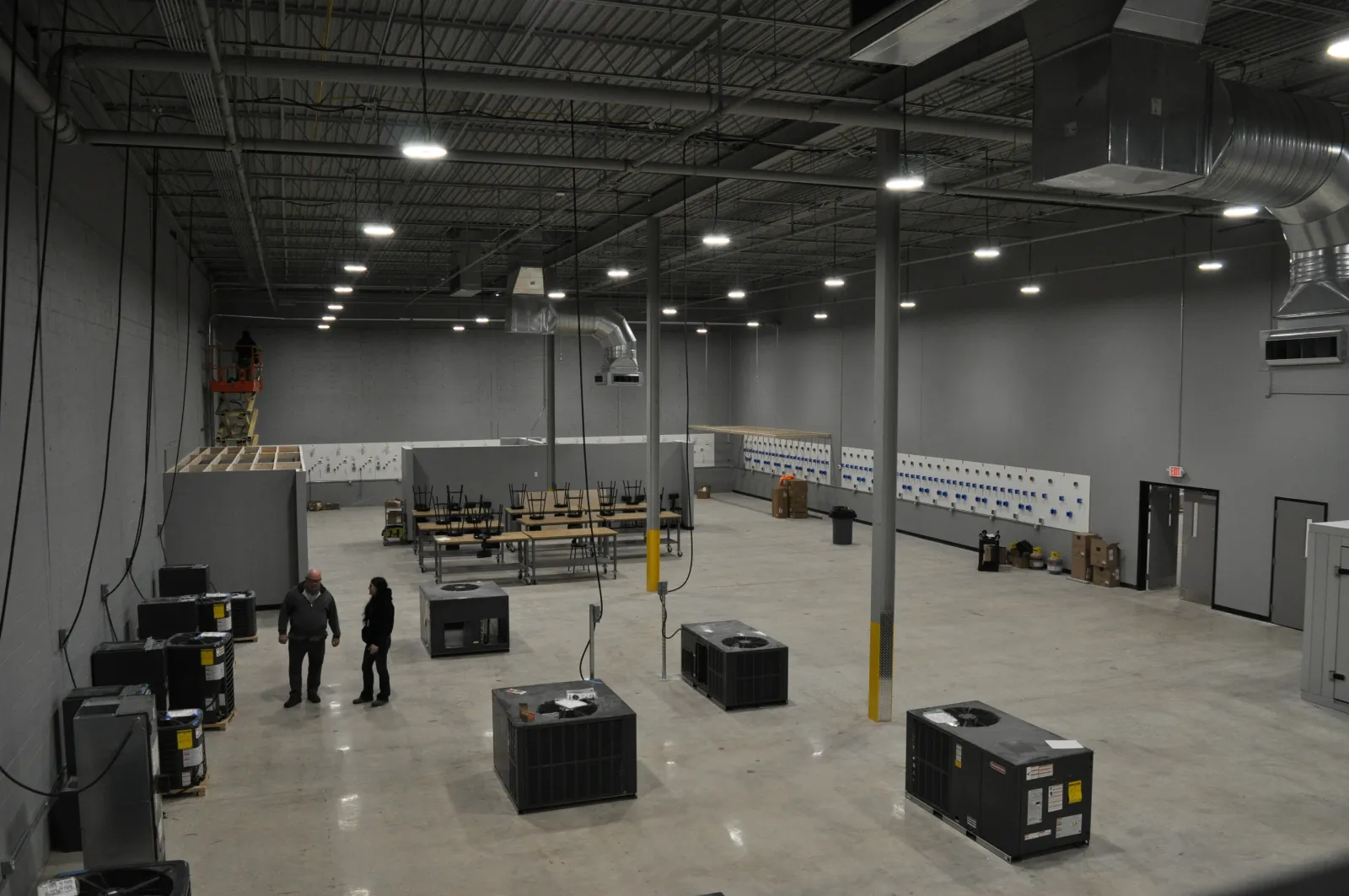 Large industrial warehouse interior with HVAC units, concrete floor, exposed ceiling, and workers inside under bright lighting