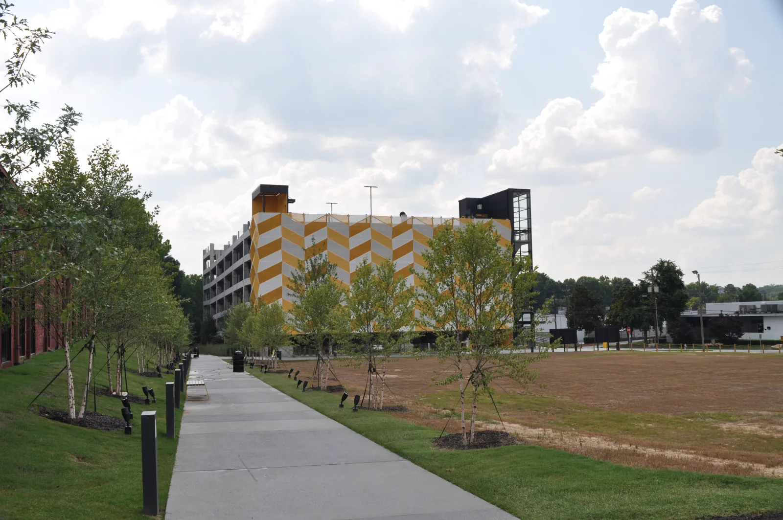 Pathway lined with young trees and lights next to a yellow and white patterned building under a cloudy sky.