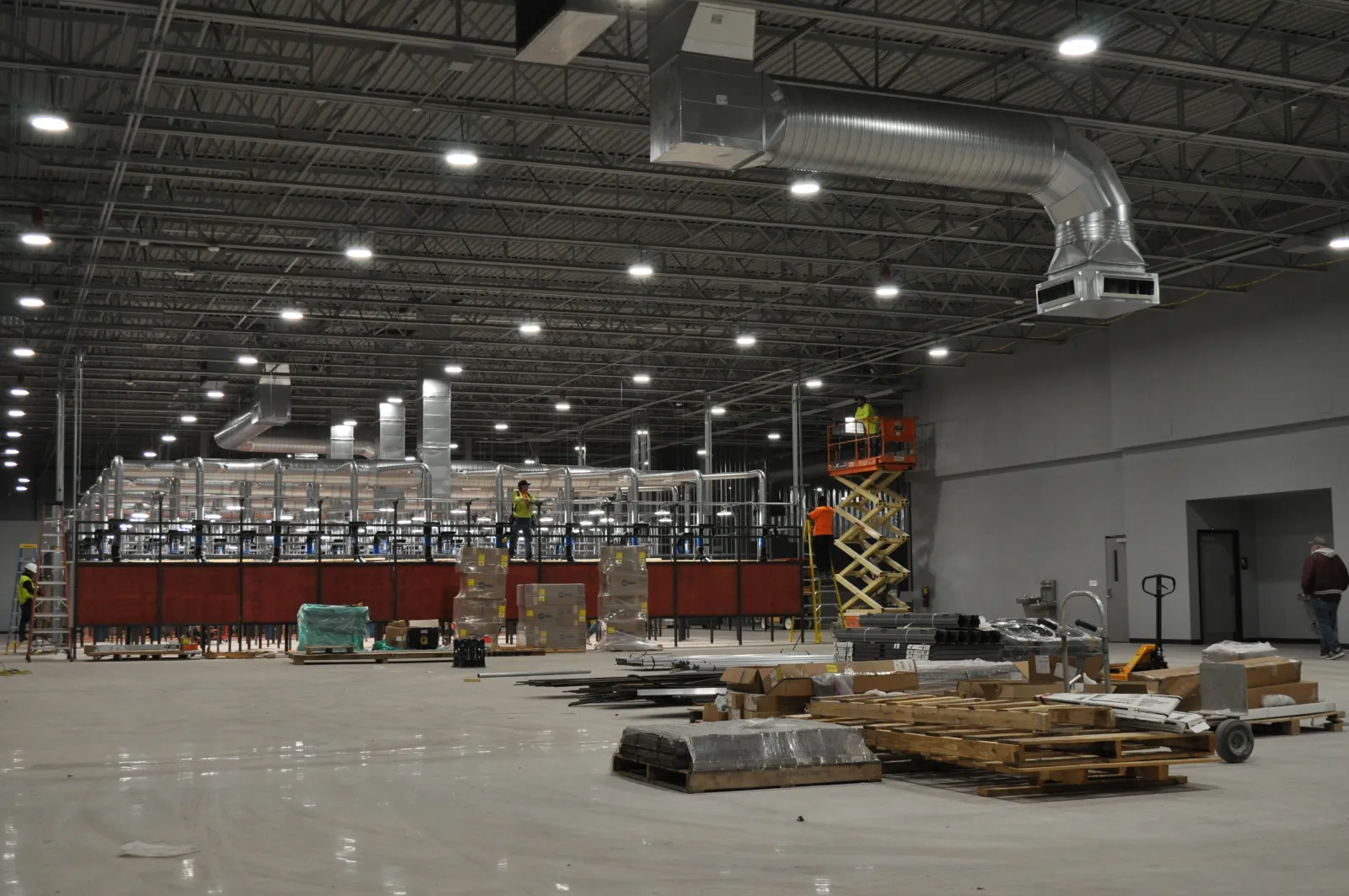 Large industrial warehouse under construction with workers, scaffolding, pallets, and HVAC ducts visible.