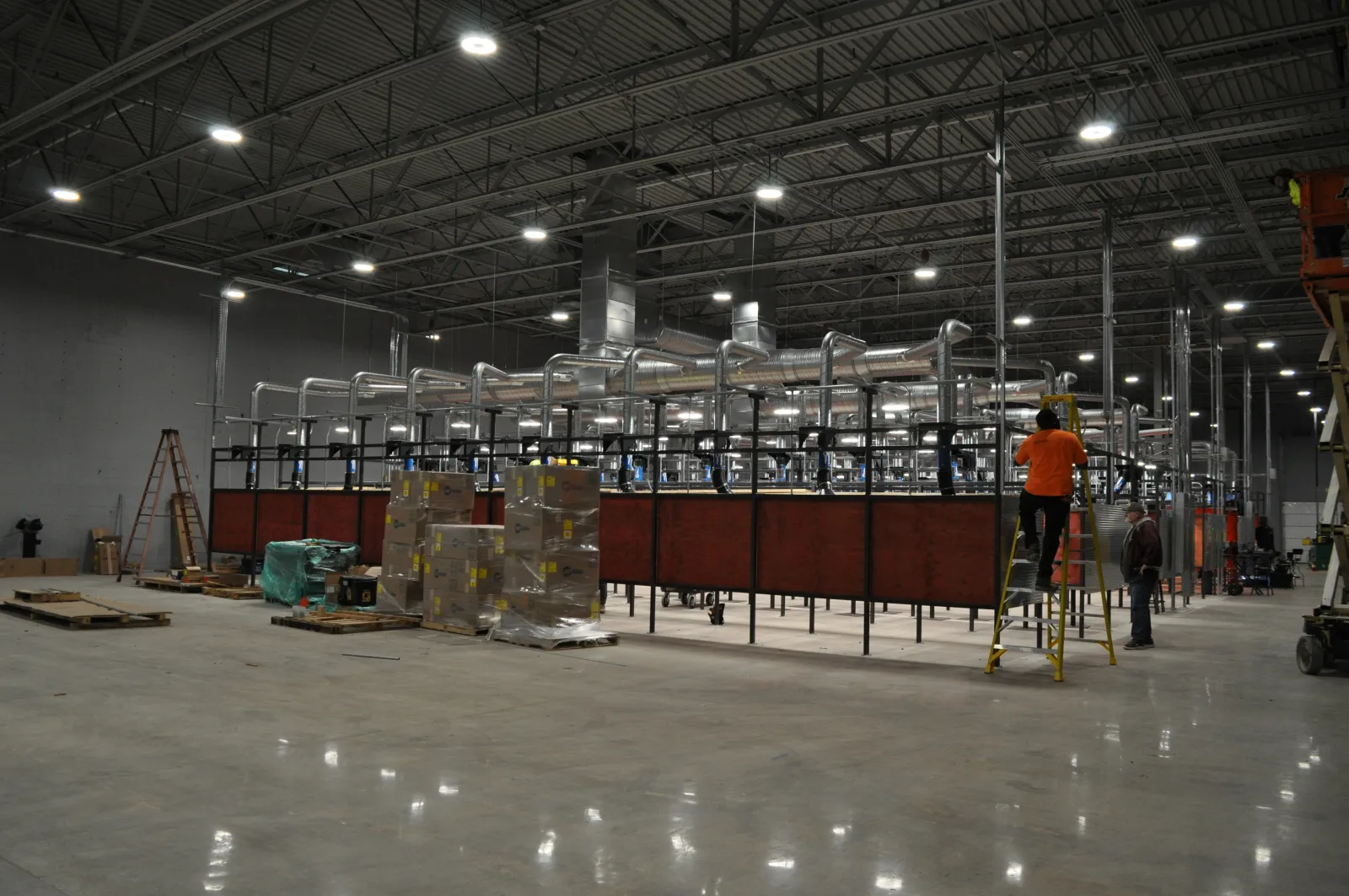 Large industrial warehouse with ventilation system, packed boxes, and workers installing equipment on a ladder.