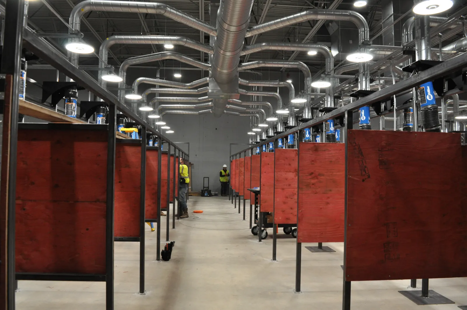 Indoor industrial workshop with rows of red wooden workstations and overhead ventilation pipes in a large space.