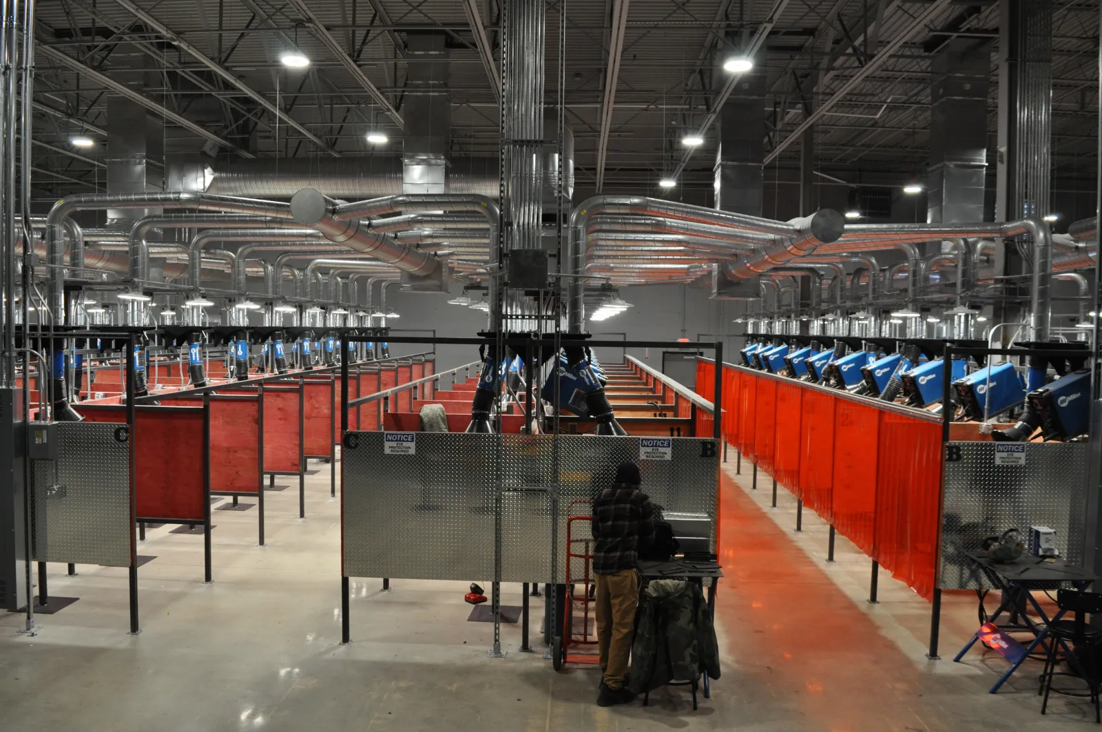 Industrial welding workshop with multiple welding stations separated by orange safety screens and equipped with blue welding machines.