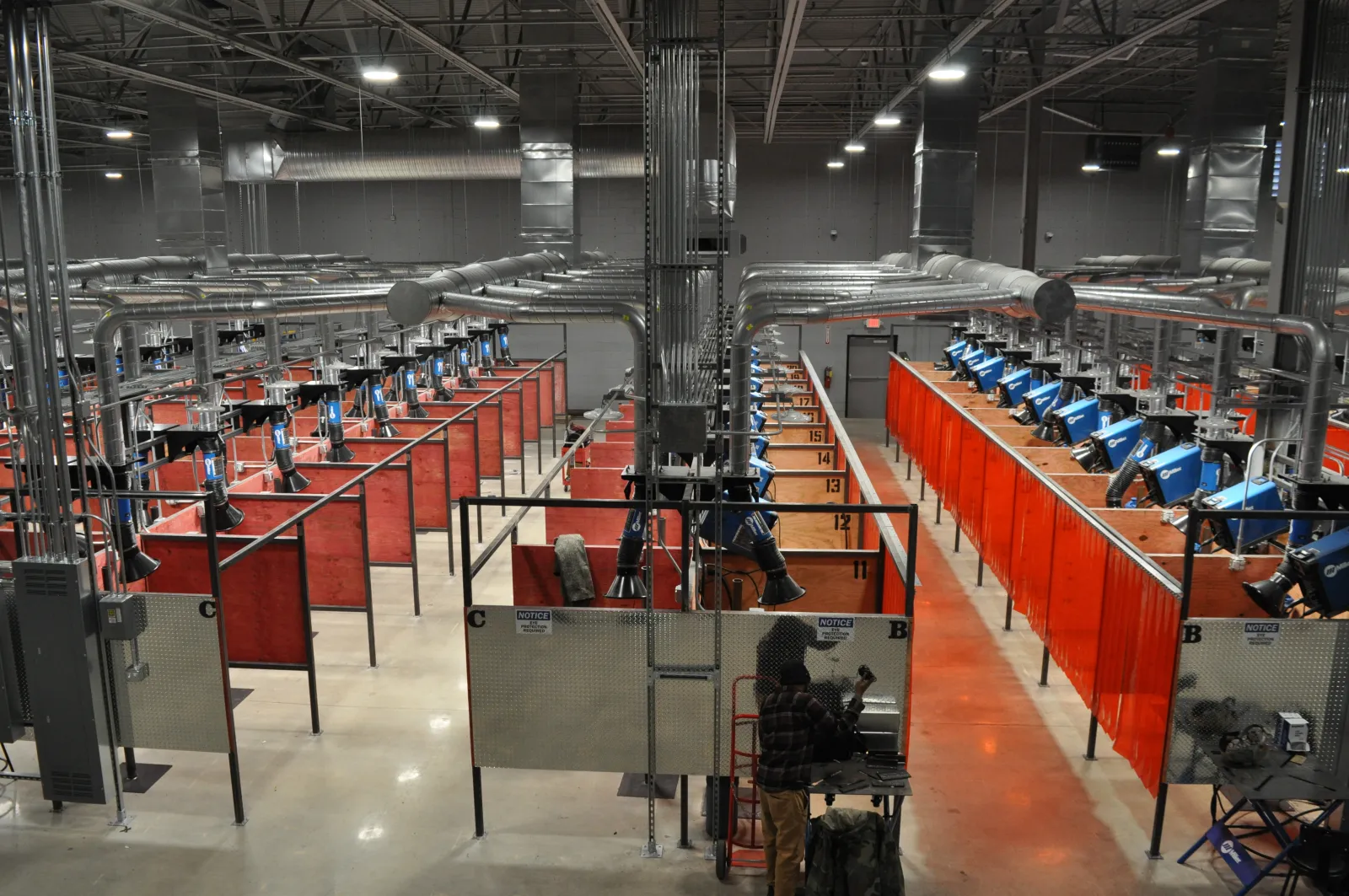 Industrial workshop with rows of welding stations separated by red and orange safety curtains under bright overhead lights.